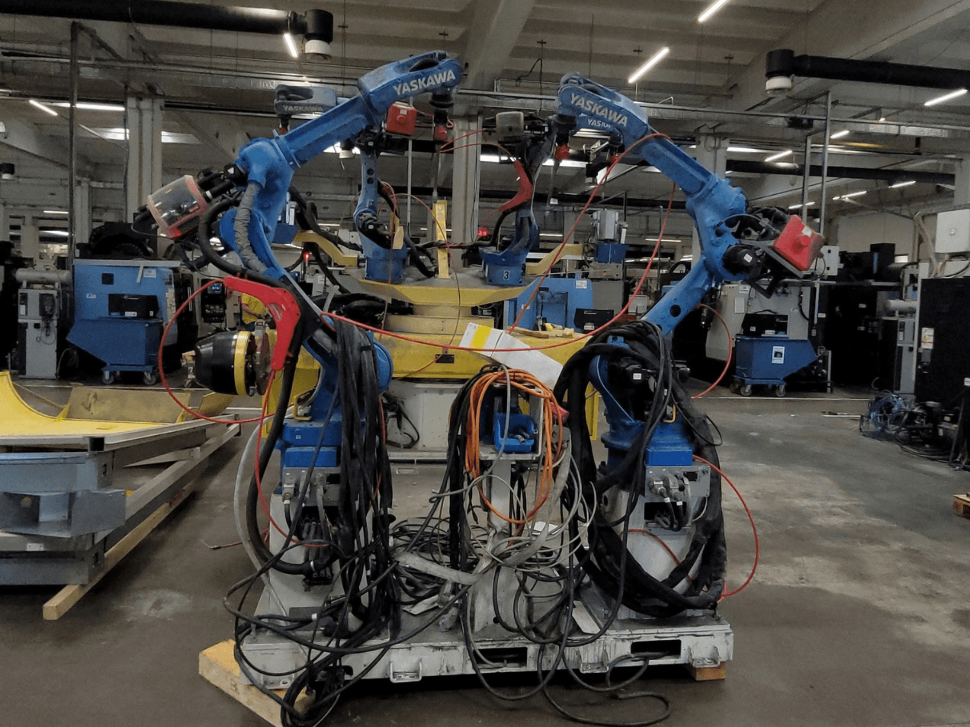 Yaskawa robotizētā metināšanas stacija RWC L1_OS 15330, skats no priekšpuses, kurā redzami divi zili roboti ar garām kabeļu vadu un metināšanas rīkiem.