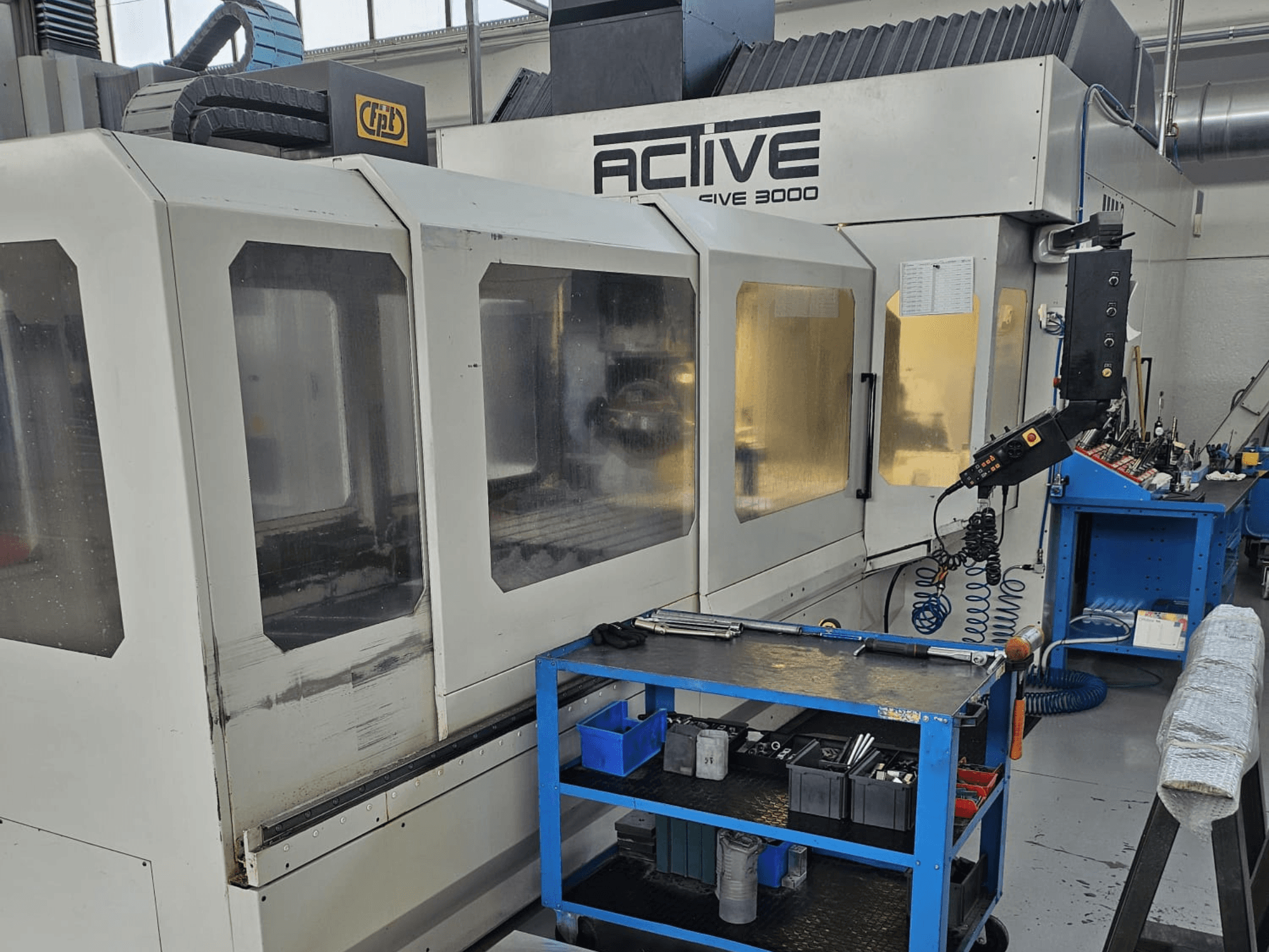 Active 3000 CNC darbnīcā, skats no priekšpuses ar instrumentiem uz zila ratiņa, redzamas vadības ierīces un darba zona darbnīcā.