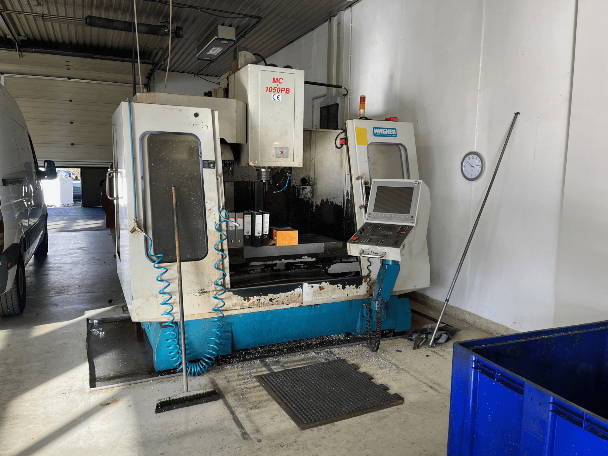 WAGNER MC 1050PB CNC darbnīcā, skats no priekšpuses, ar vadības paneli un darba zonu; redzama zila gaisa šļūtene un tīrīšanas instrumenti.