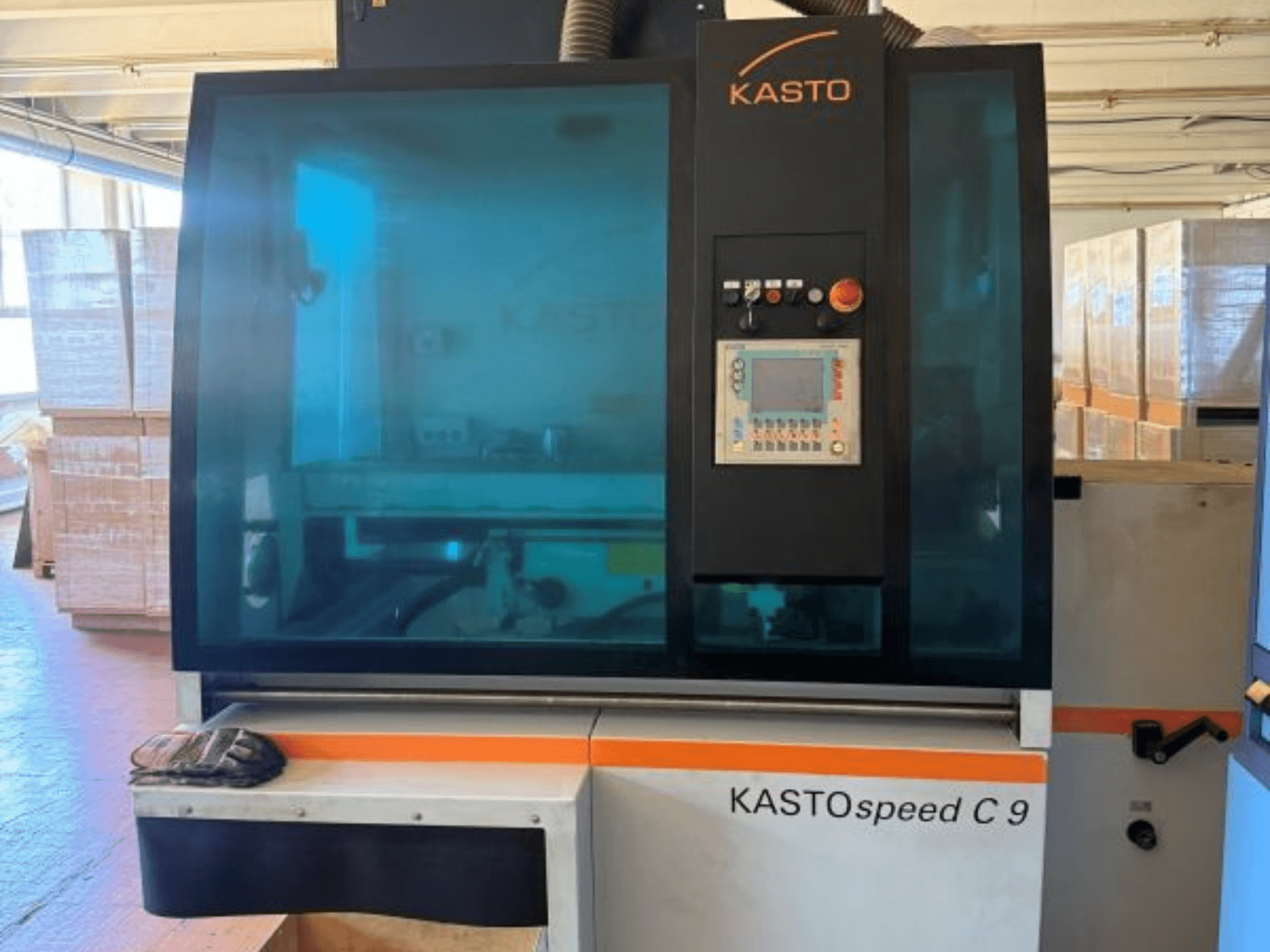 KASTO KASTOspeed C 9 metāla ripzāģa skats no priekšpuses, kurā redzams vadības panelis un drošības stikls, fonā — koka kastes.