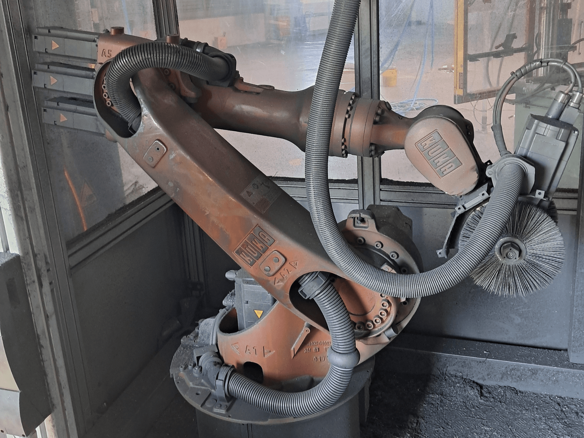 KUKA KR 16-2 robota roka, skatīta no sāniem, kurā redzami vadi, sukas un mehāniskie savienojumi, novietota darba vidē.
