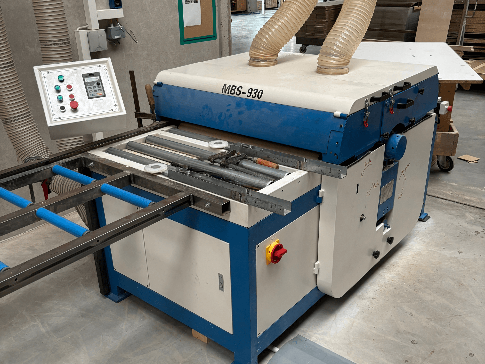 MBS-930 kokapstrādes mašīna ar vadības paneli un veltņiem, skatoties no sāniem, ar putekļu nosūces šļūtenēm.