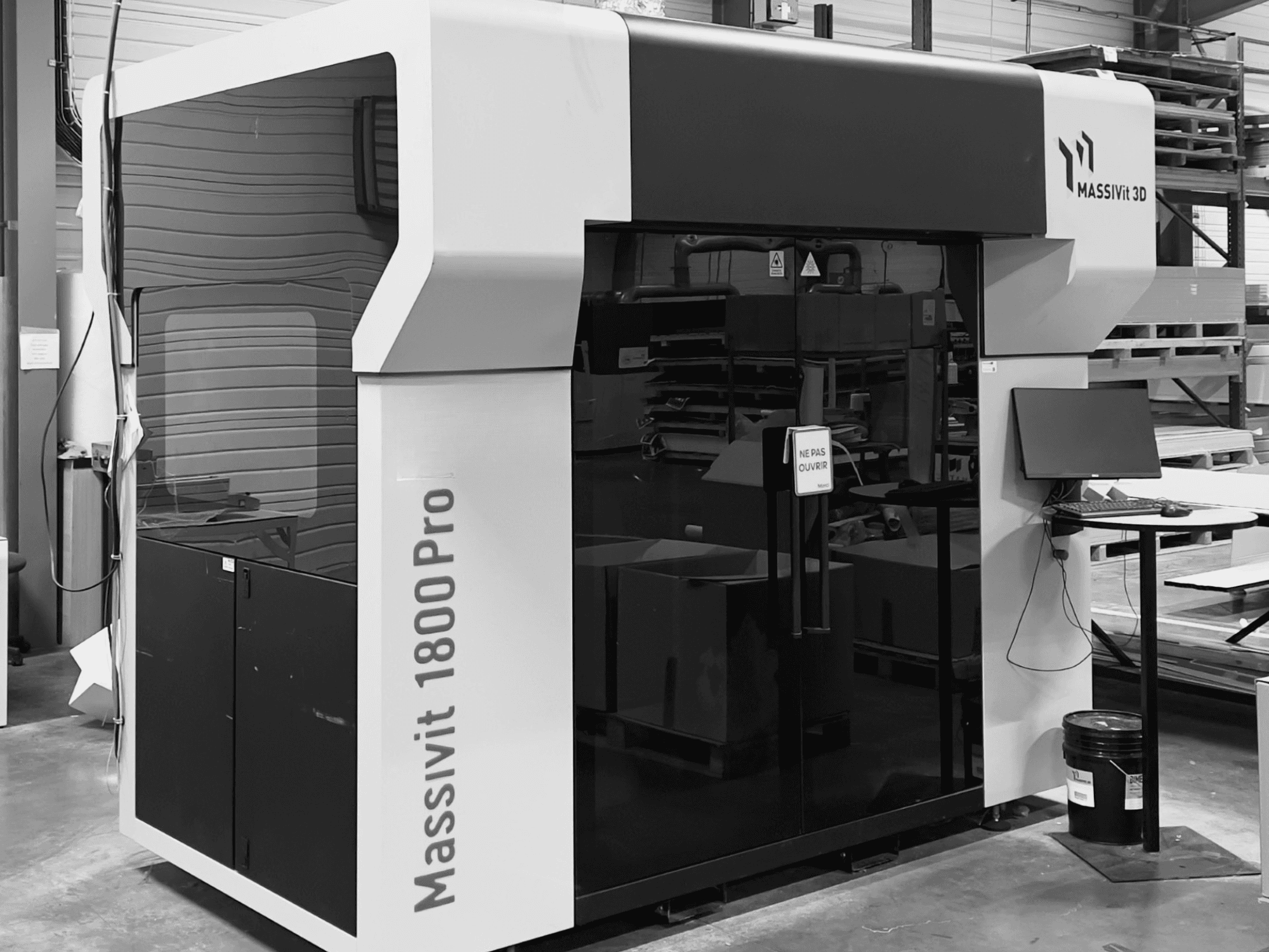 Plastmasas 3D printeris MASSIVIT 1800 PRO darbnīcā, skats no priekšpuses, kurā redzama datora darba vieta un vadības panelis.