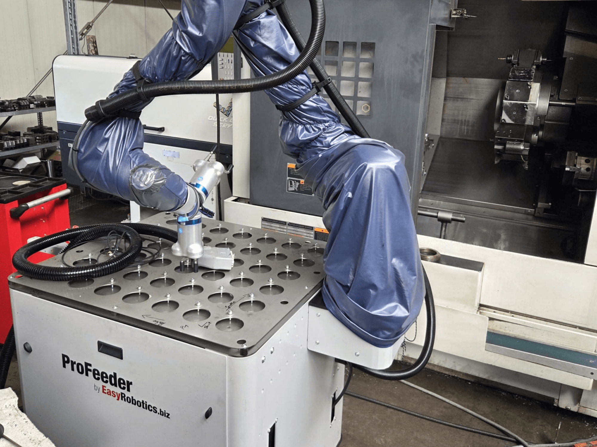 Sadarbības robots EASYROBOTICS ProFeeder no priekšpuses, ar robota roku, kas novietota virs perforēta metāla galda.