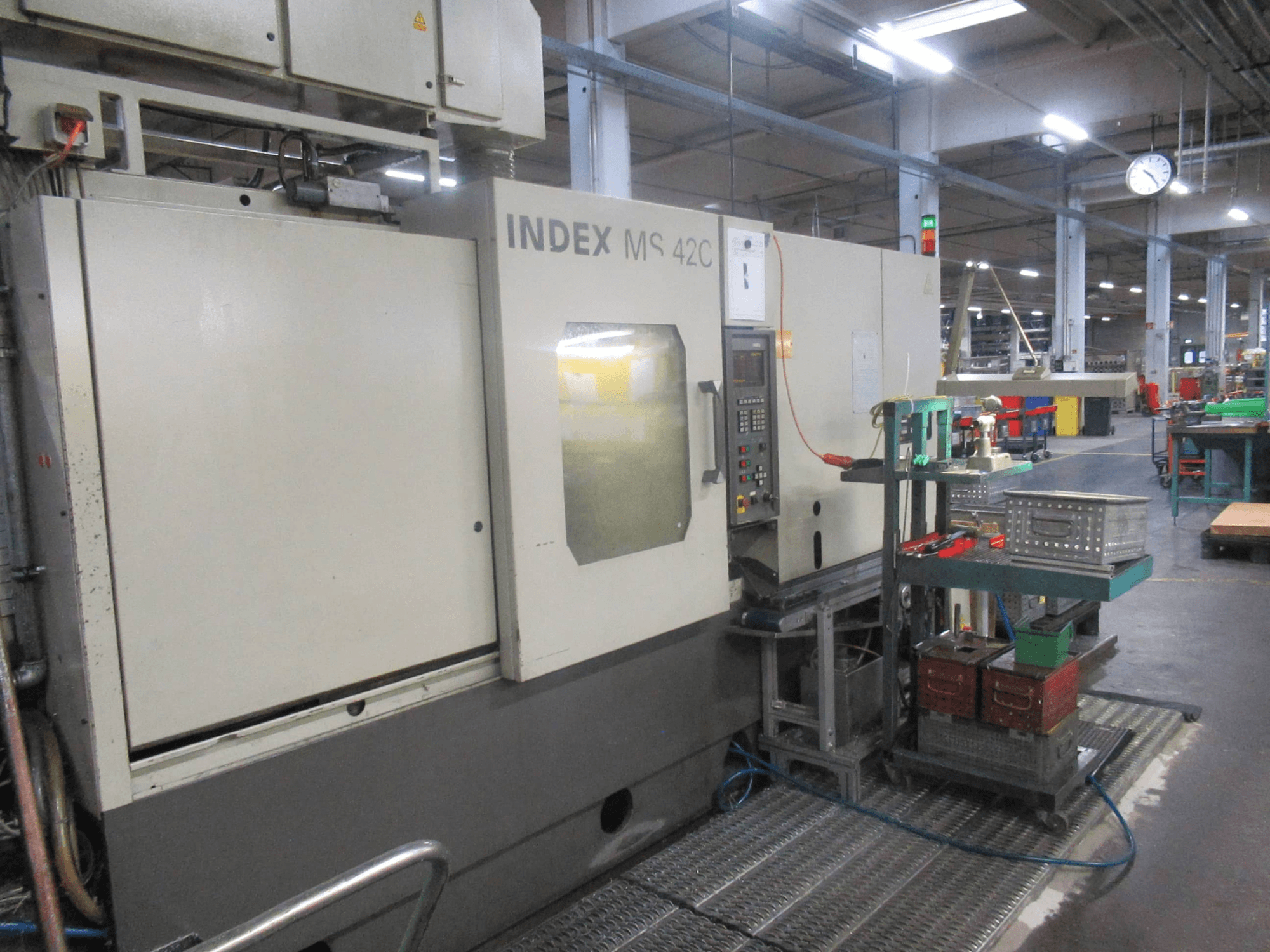 INDEX MS 42C CNC darbnīcā, skats no sāniem, kurā redzams vadības panelis un instrumentu glabāšanas vieta uz riteņu ratiņiem.