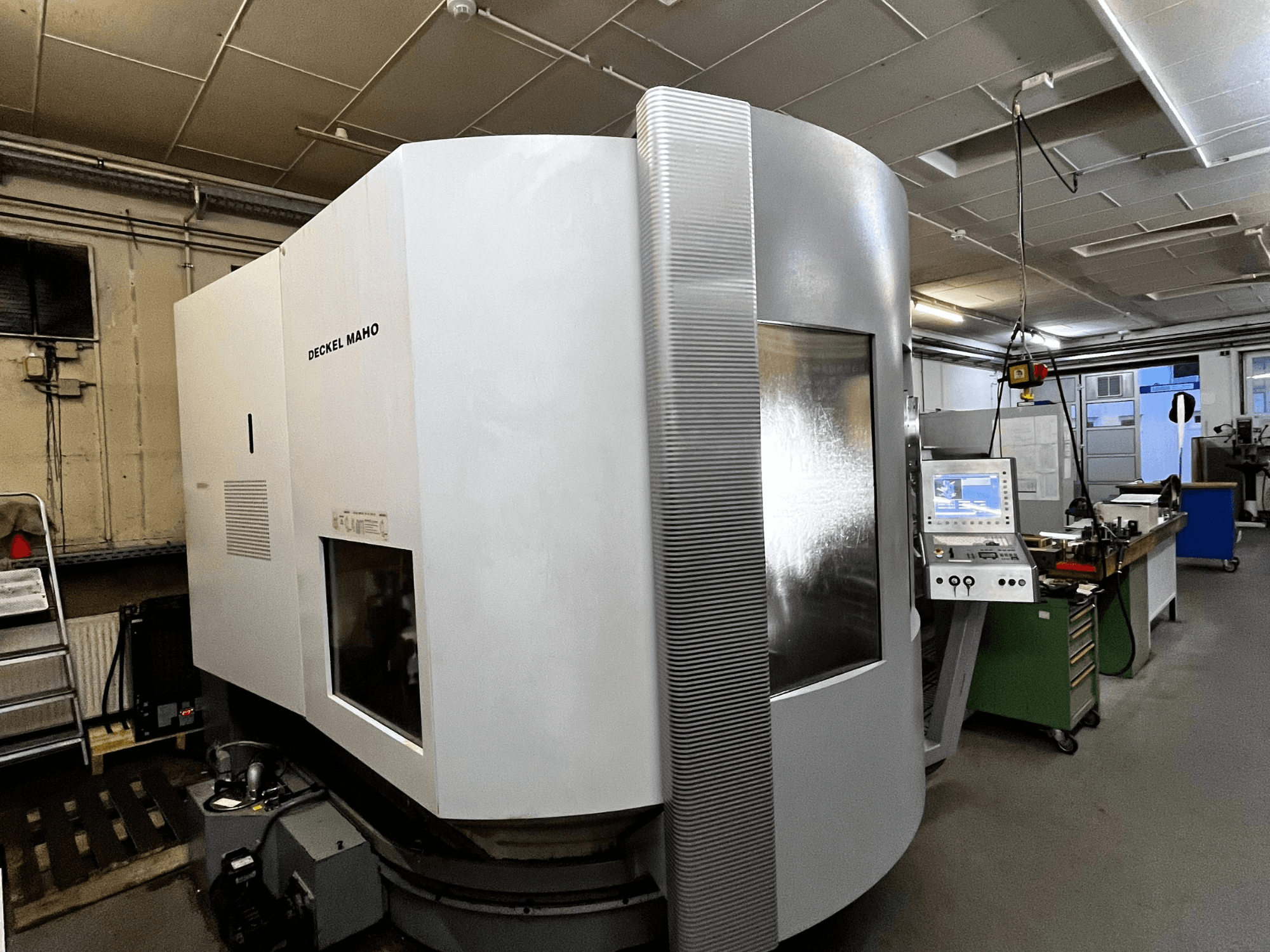 Deckel Maho CNC mašīna, skats no priekšpuses, balta ārpuse, redzams vadības panelis, darbnīcas iestatījumi.