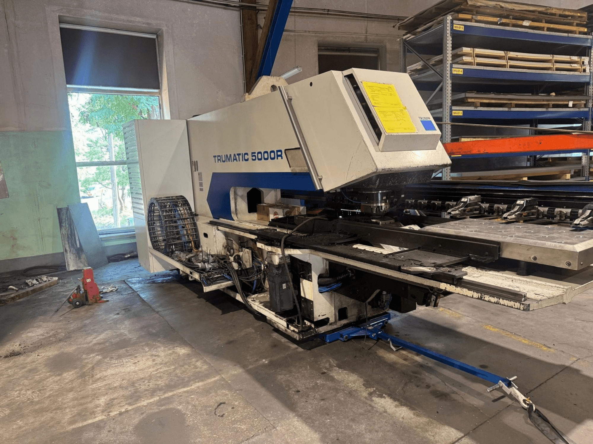 CNC perforācijas mašīna TRUMPF TRUMATIC 5000R darbnīcā, skats no priekšpuses, kurā redzamas tās vadības ierīces un perforācijas zona.