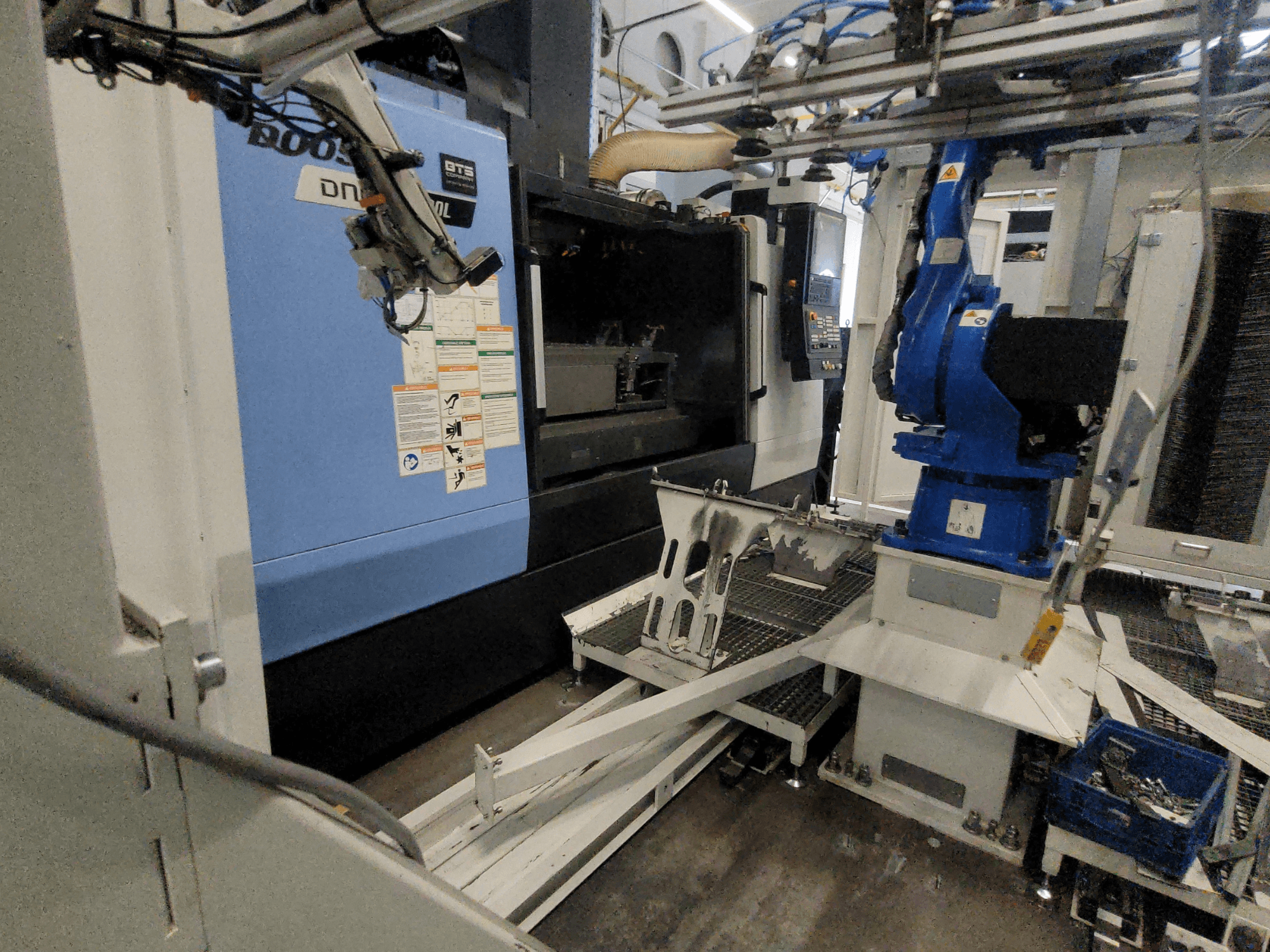 DOOSAN DNM 6700L vertikālā apstrādes centra sānskats, kurā redzama robota roka un vadības panelis, parādot iekārtas konfigurāciju.
