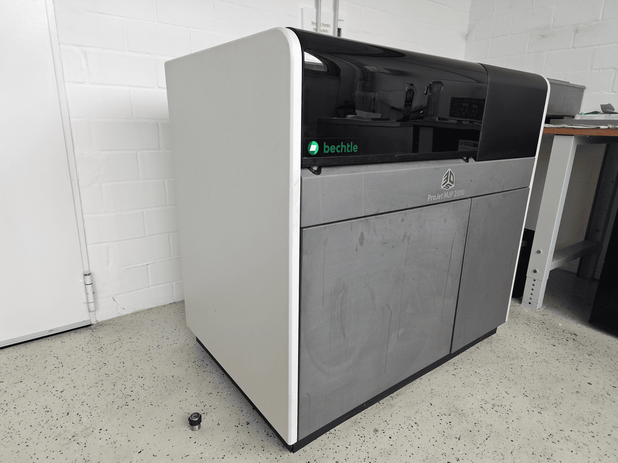 3D Systems ProJet MJP 2500 Plus plastmasas printeris, skats no priekšpuses; tam ir elegants dizains ar tumšu augšdaļu un gaišu korpusu.