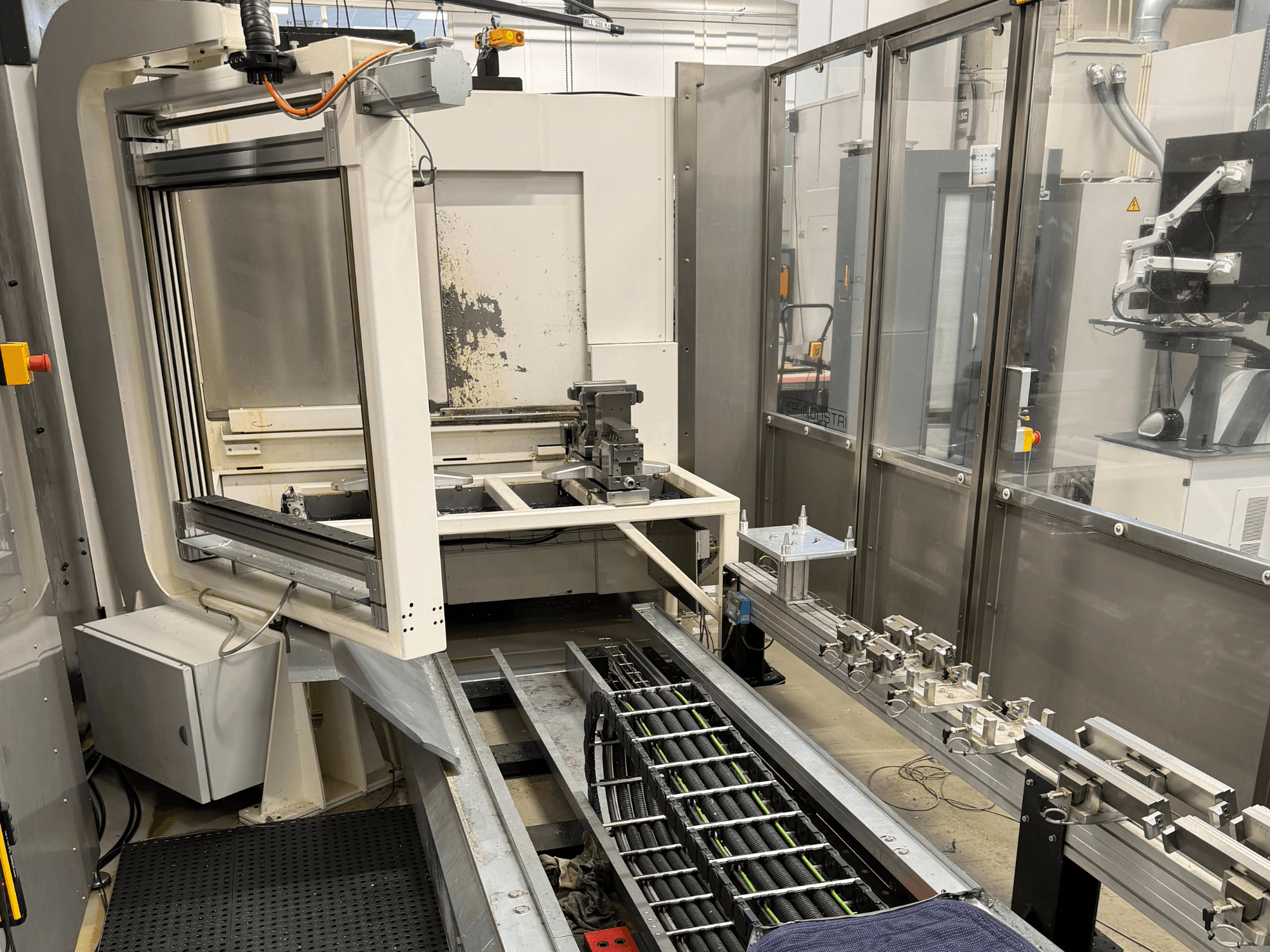 Automatizēta rūpnieciskā darbstacija ar robotizētu roku, sliežu sistēmu un vadības paneli sānu skatā.