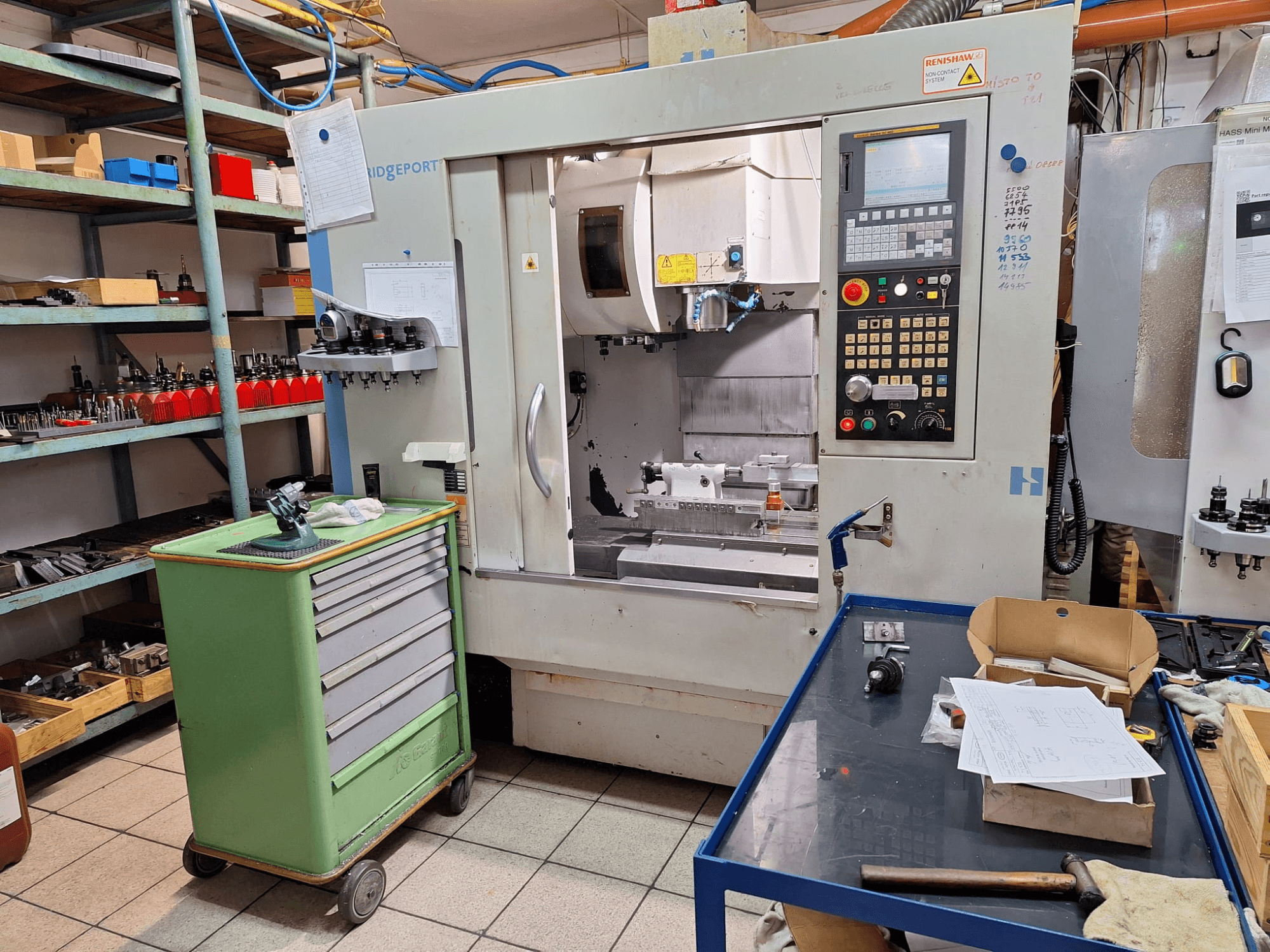 Hardinge Bridgeport GX 710 vertikālā apstrādes centra priekšskats ar vadības pulti, instrumentiem un plauktiem darbnīcas vidē.