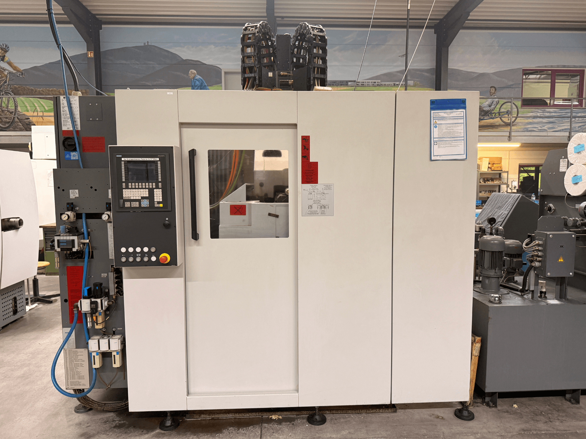 Balts CNC apstrādes centrs ar vadības paneli, skats no priekšpuses, ar pogām un digitālo displeju, darbnīcā.