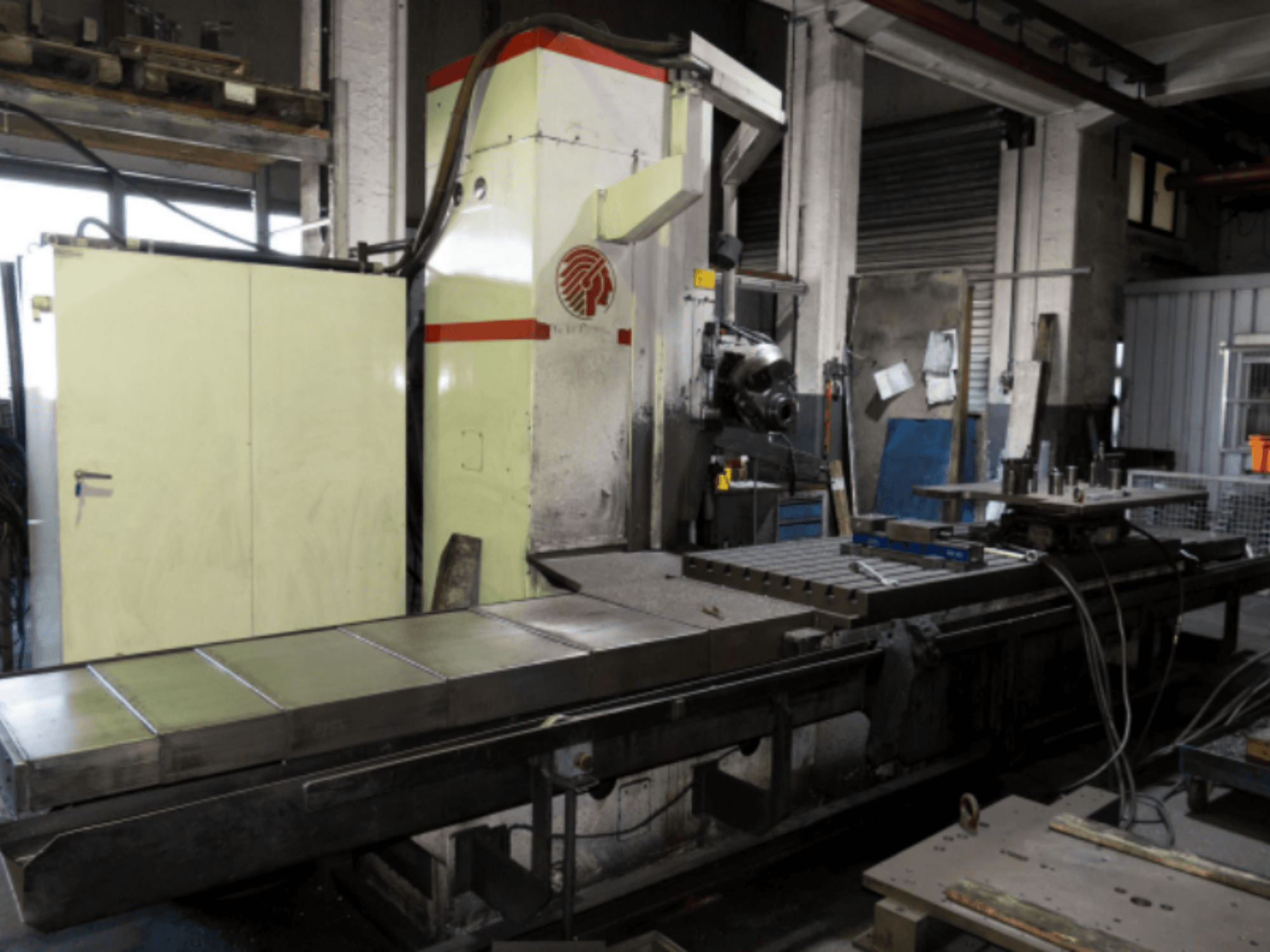 Rūpnieciskā frēzmašīna ar lielu galdu, skats no sāniem, ar vadības ierīcēm un stiprinājumiem darbnīcā.