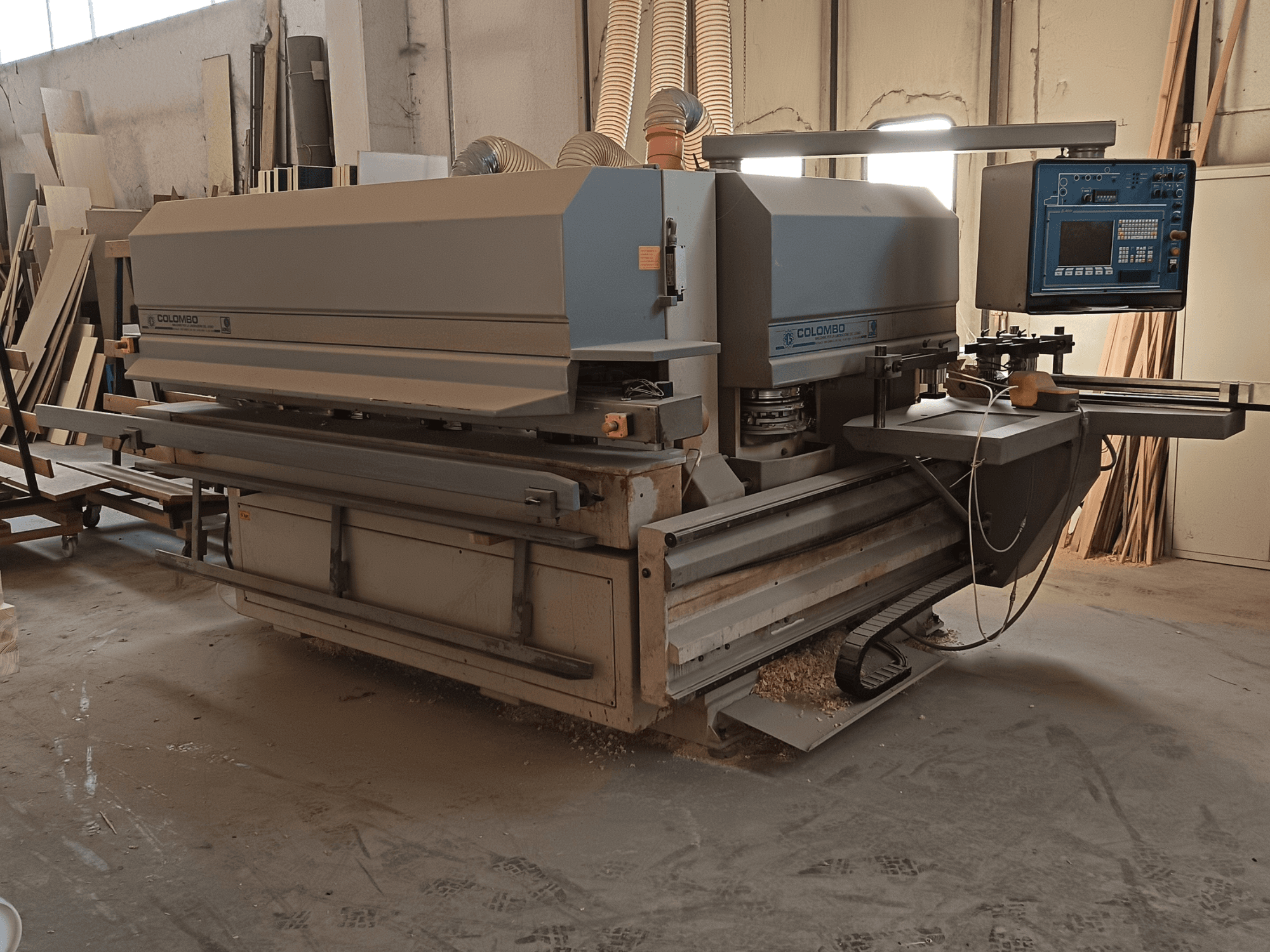 Colombo kokapstrādes mašīna, skats no sāniem, kurā redzams vadības panelis, griešanas asmeņi un zāģa putekļu uzkrāšanās darbnīcā.