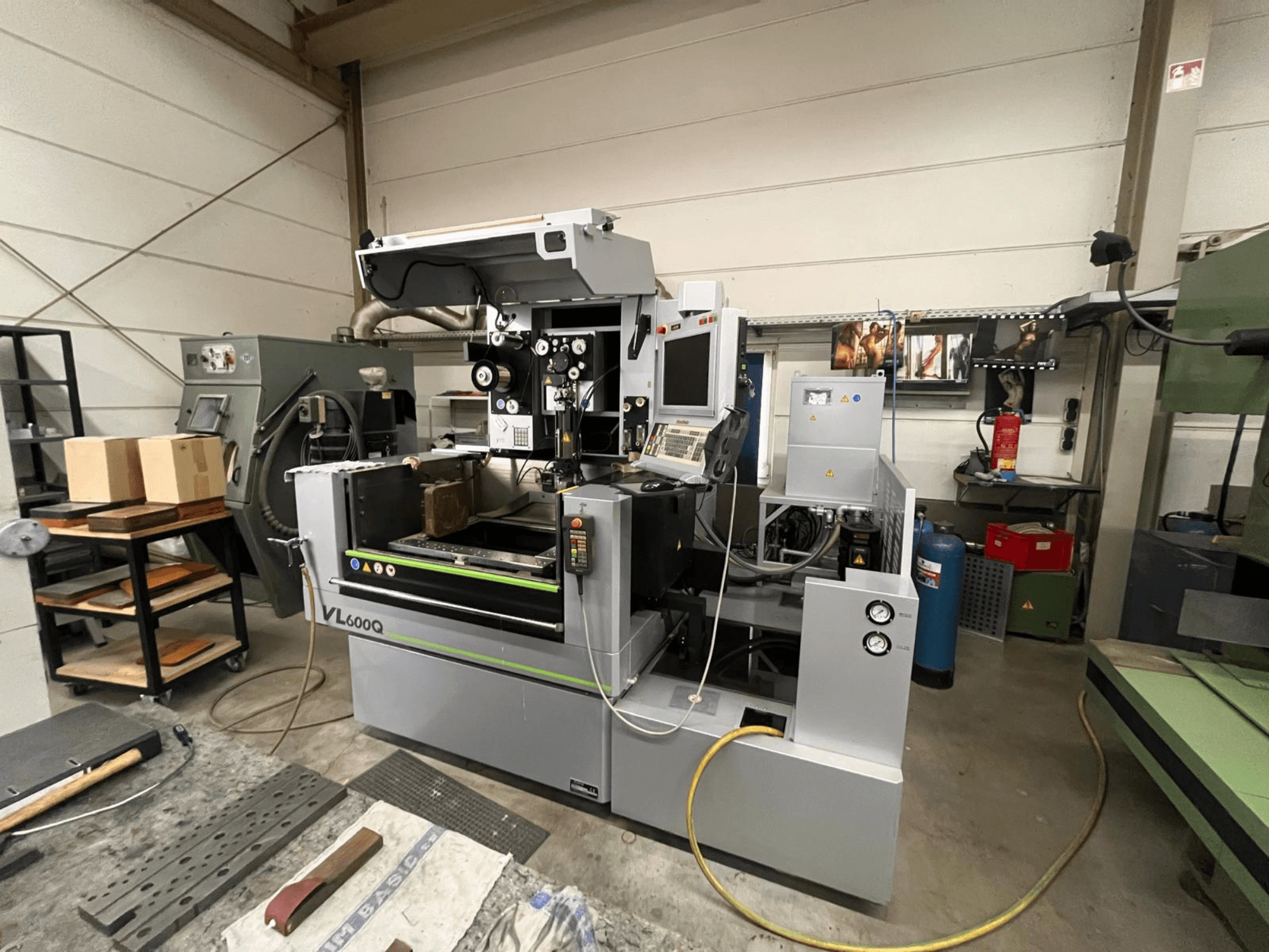 VL600Q CNC darbnīcā, skats no priekšpuses, ar vadības paneli, novietotu materiālu un dažādiem instrumentiem blakus.