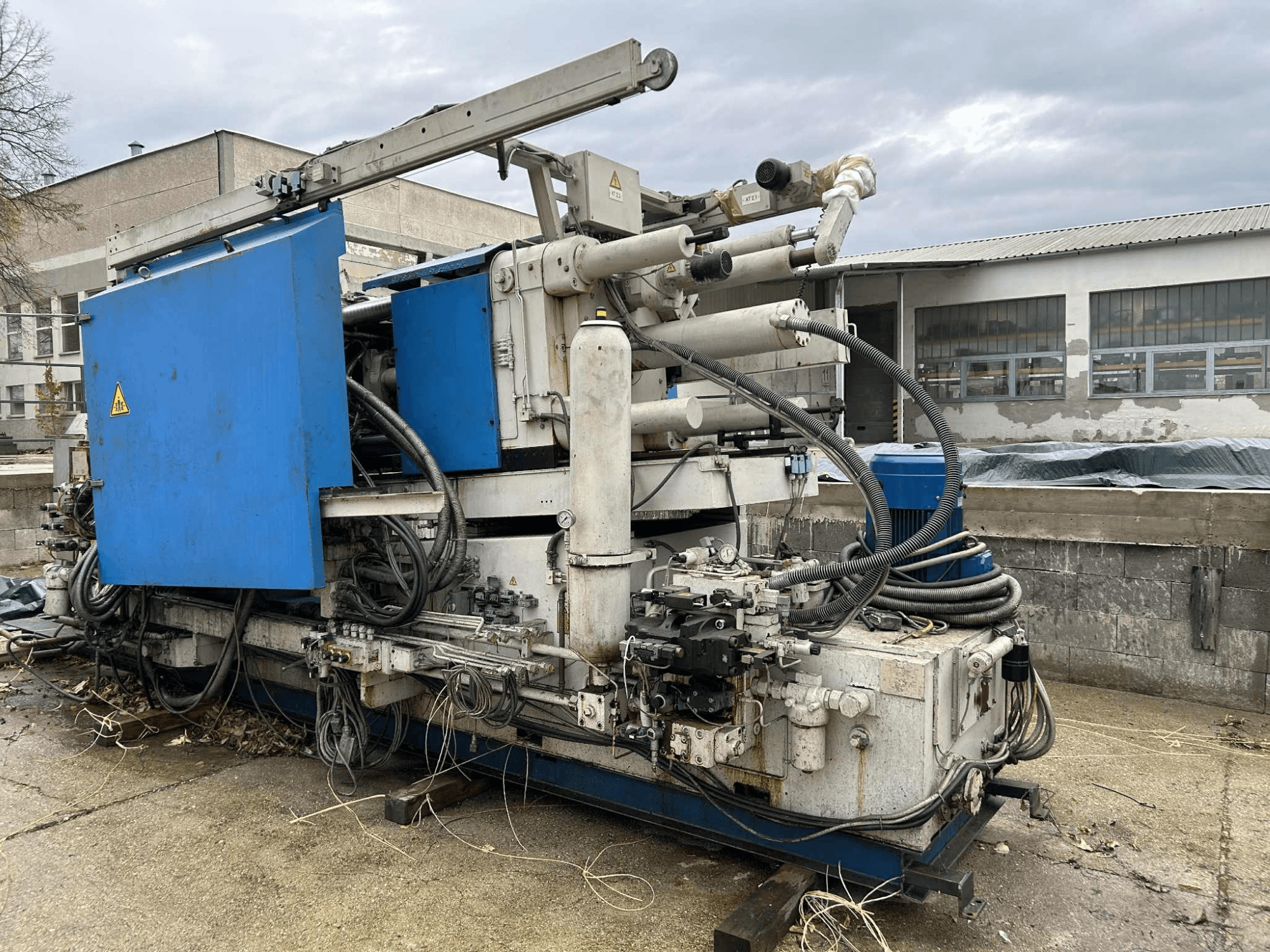 Liela rūpnieciska hidrauliskā mašīna ar zilu korpusu, dažādām šļūtenēm un metāla detaļām, skatoties no sāniem.