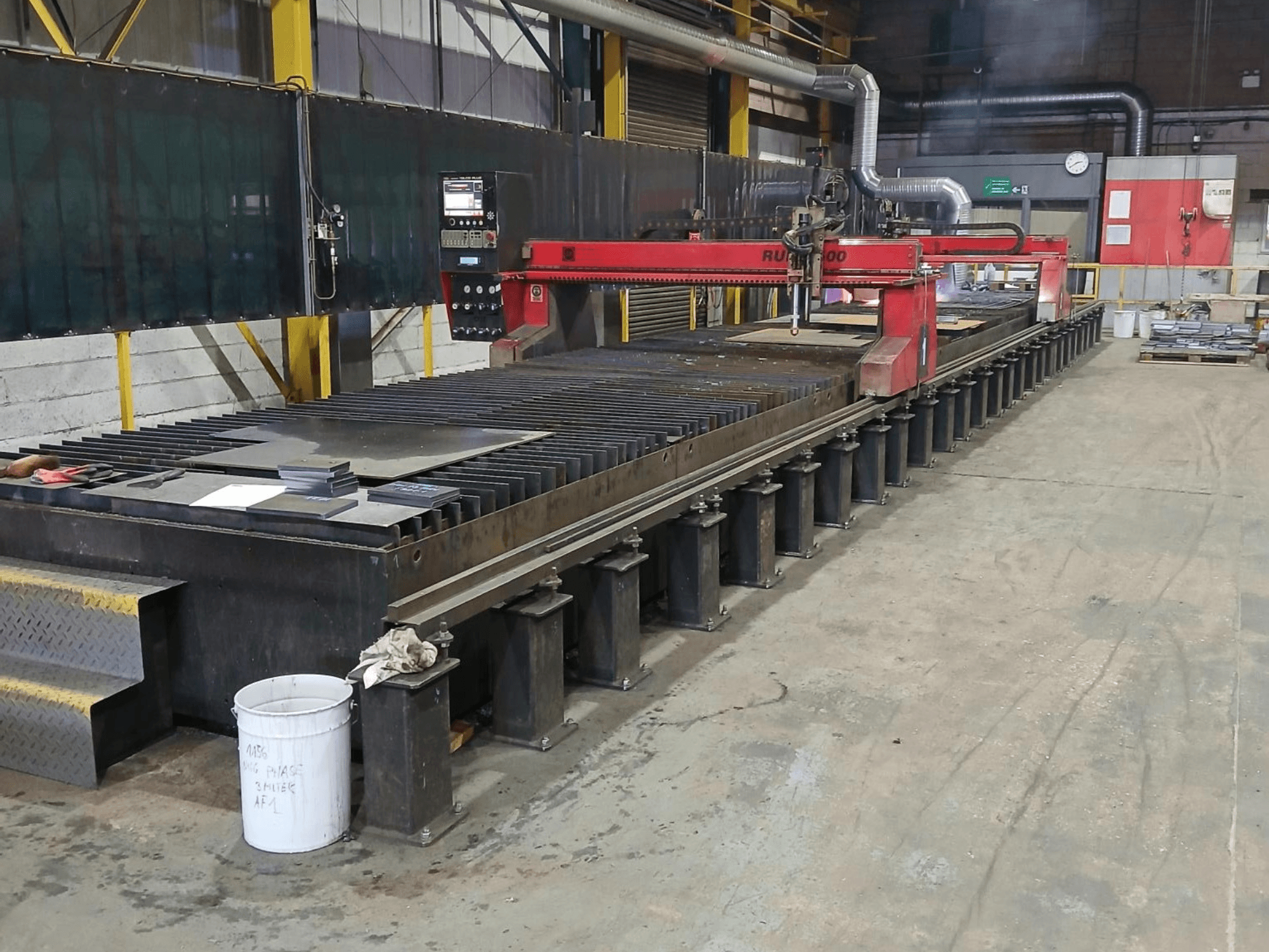 Sarkana RUIDA 300 griešanas iekārta, skats no sāniem, ar metāla loksni uz tērauda galda, kas atrodas rūpnieciskā darba telpā.