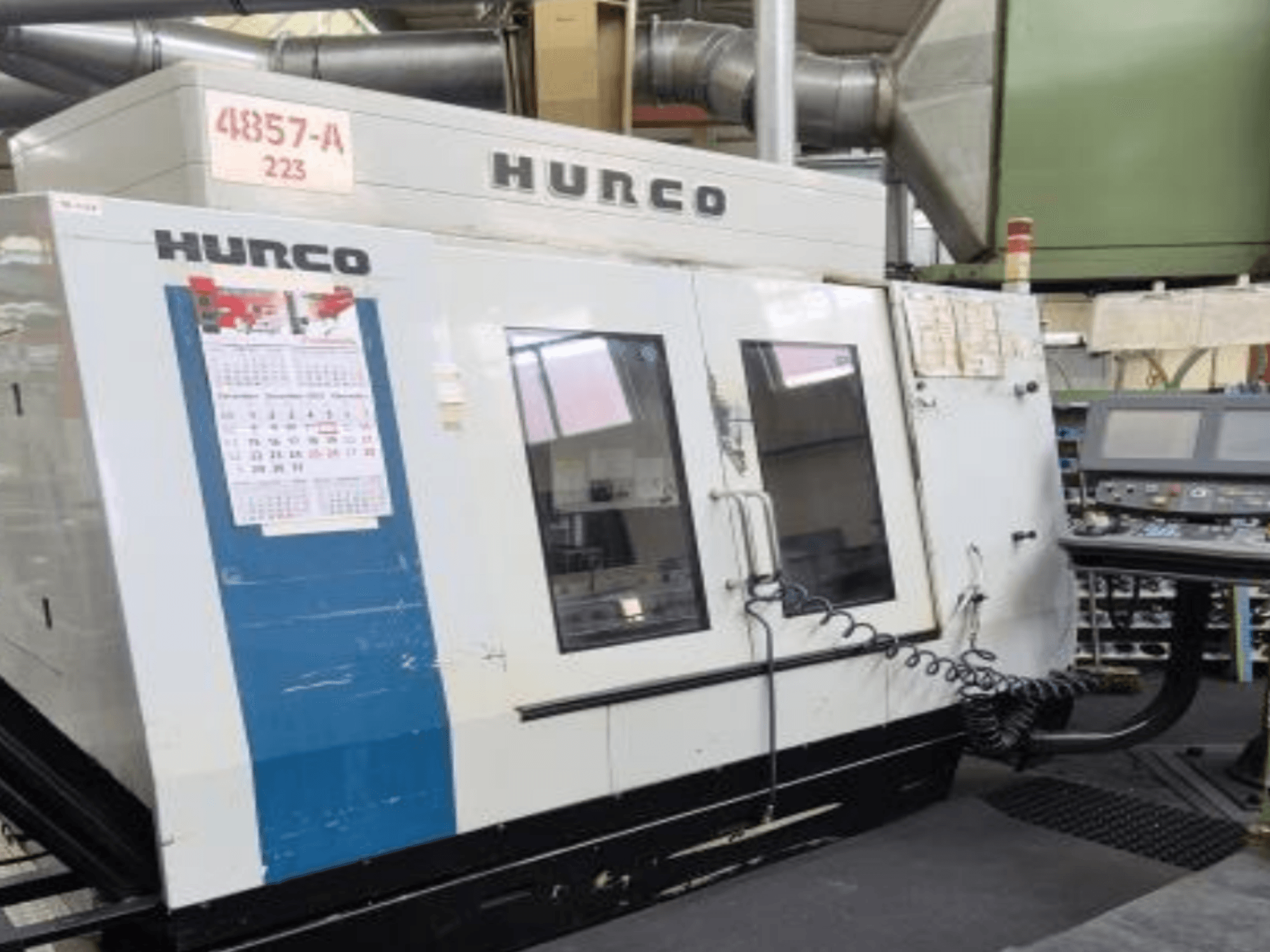 Hurco VMX 42 vertikālais apstrādes centrs priekšpusē, ar vadības paneli, kalendāru un metāla ārējām detaļām.