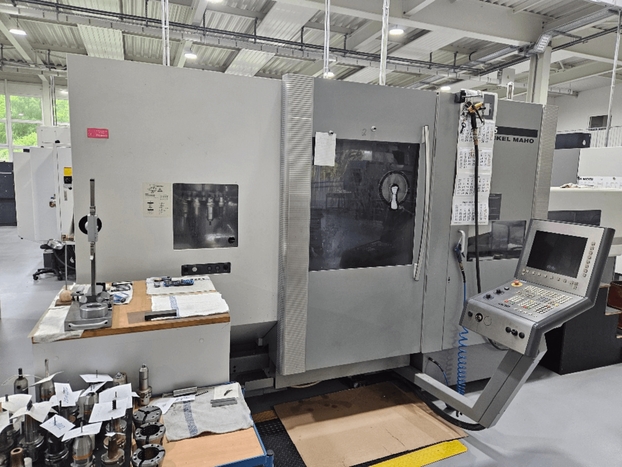 DMG Mori DMU 70 mašīnas priekšējais skats ar vadības paneli, instrumentiem uz koka galda un dažādām detaļām blakus.