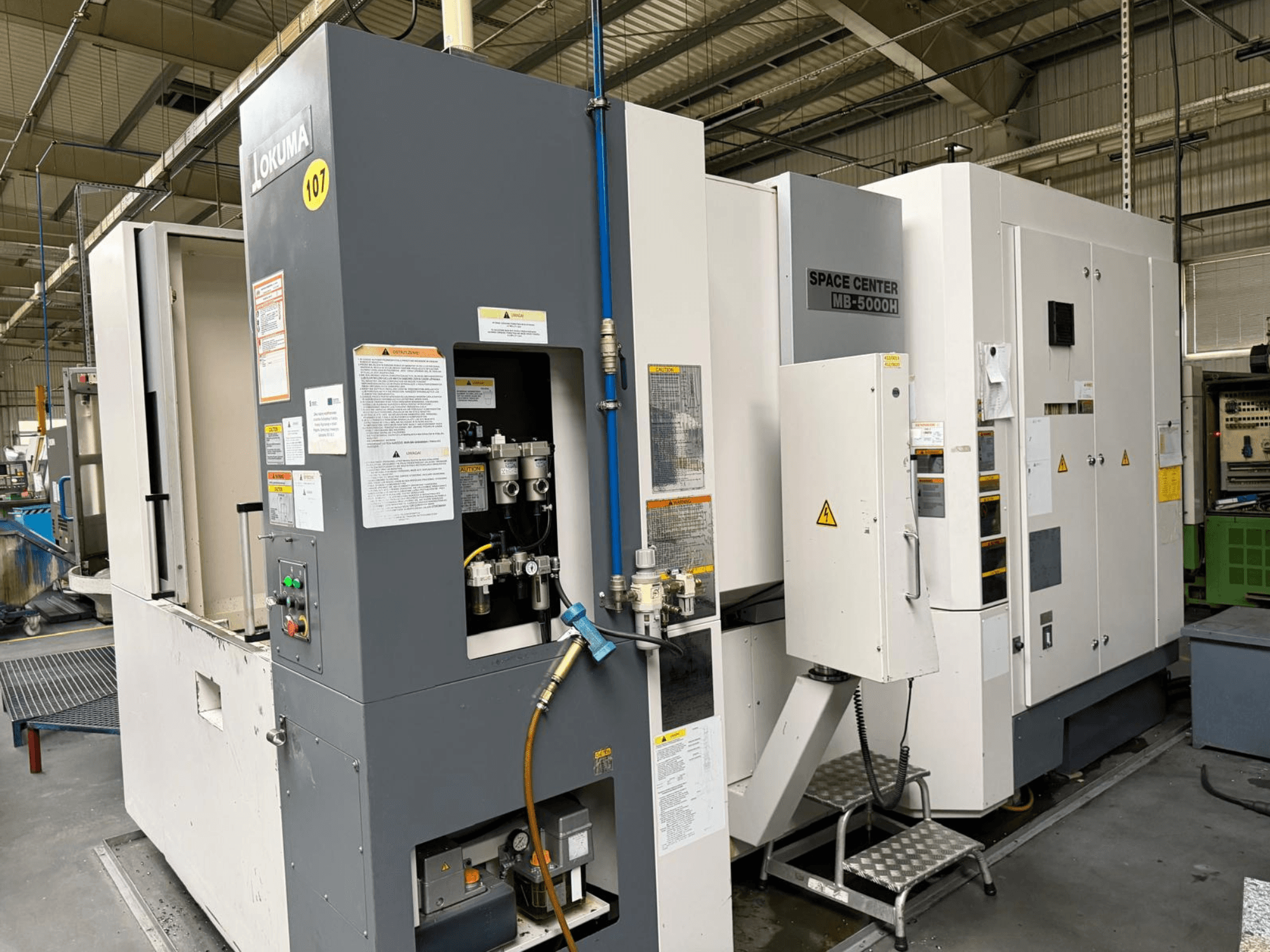 Okuma MB 5000H horizontālais apstrādes centrs, sānu skats, kurā redzams vadības panelis un mašīnu komponenti rūpnīcā.