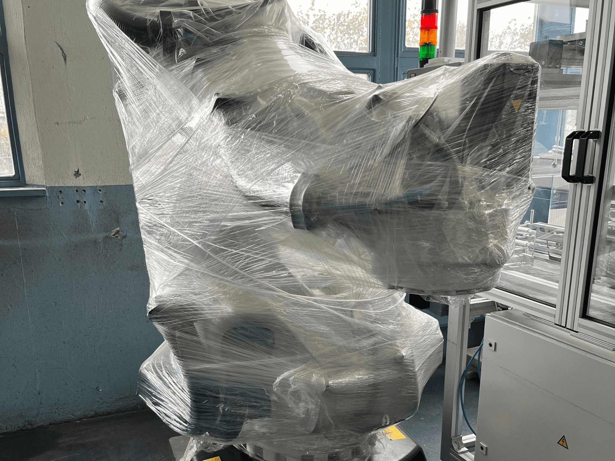 Plastmasā ietīta rūpnieciskā robota roka, skats no priekšpuses. Ietver vadības bloku un indikatoru. Zīmols nav redzams.