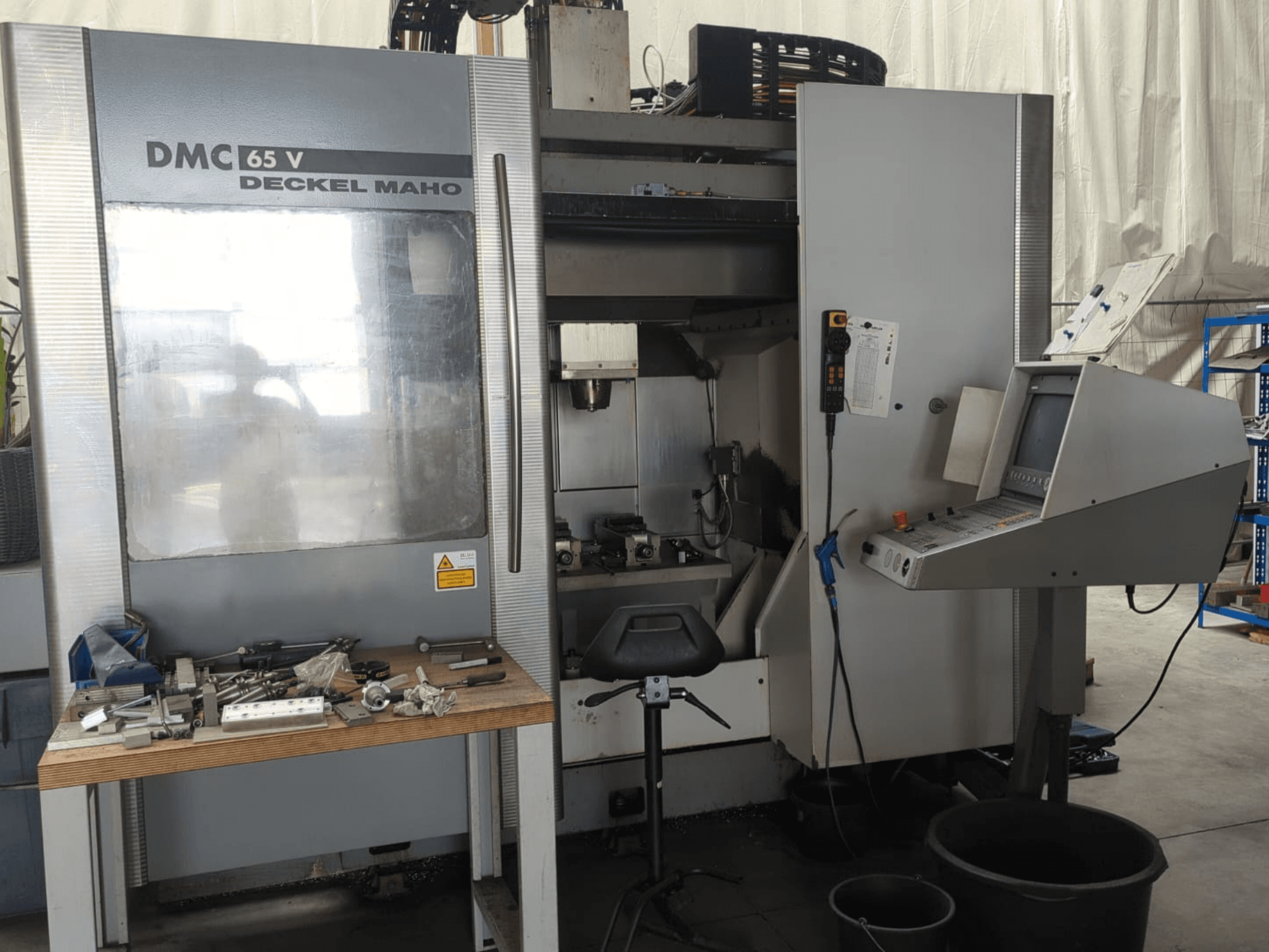 DECKEL MAHO DMC 65 V vertikālā apstrādes centra skats no priekšpuses ar vadības paneli, instrumentiem uz galda un darba zonas konfigurāciju.