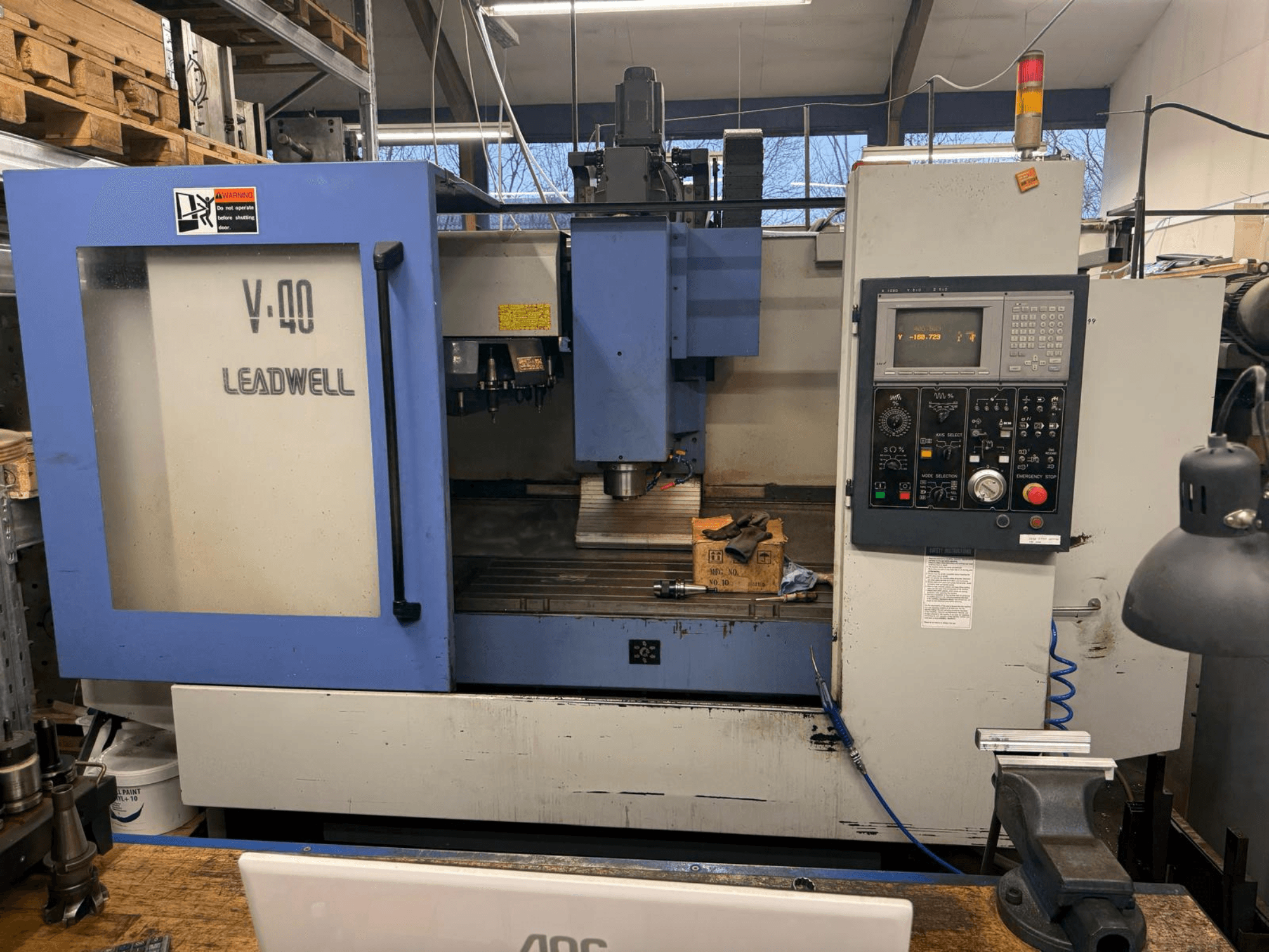 LEADWELL V-40 CNC frēzmašīna, skats no priekšpuses, ar vadības paneli, griezējinstrumentiem un skrūvgriezi uz darba galda.
