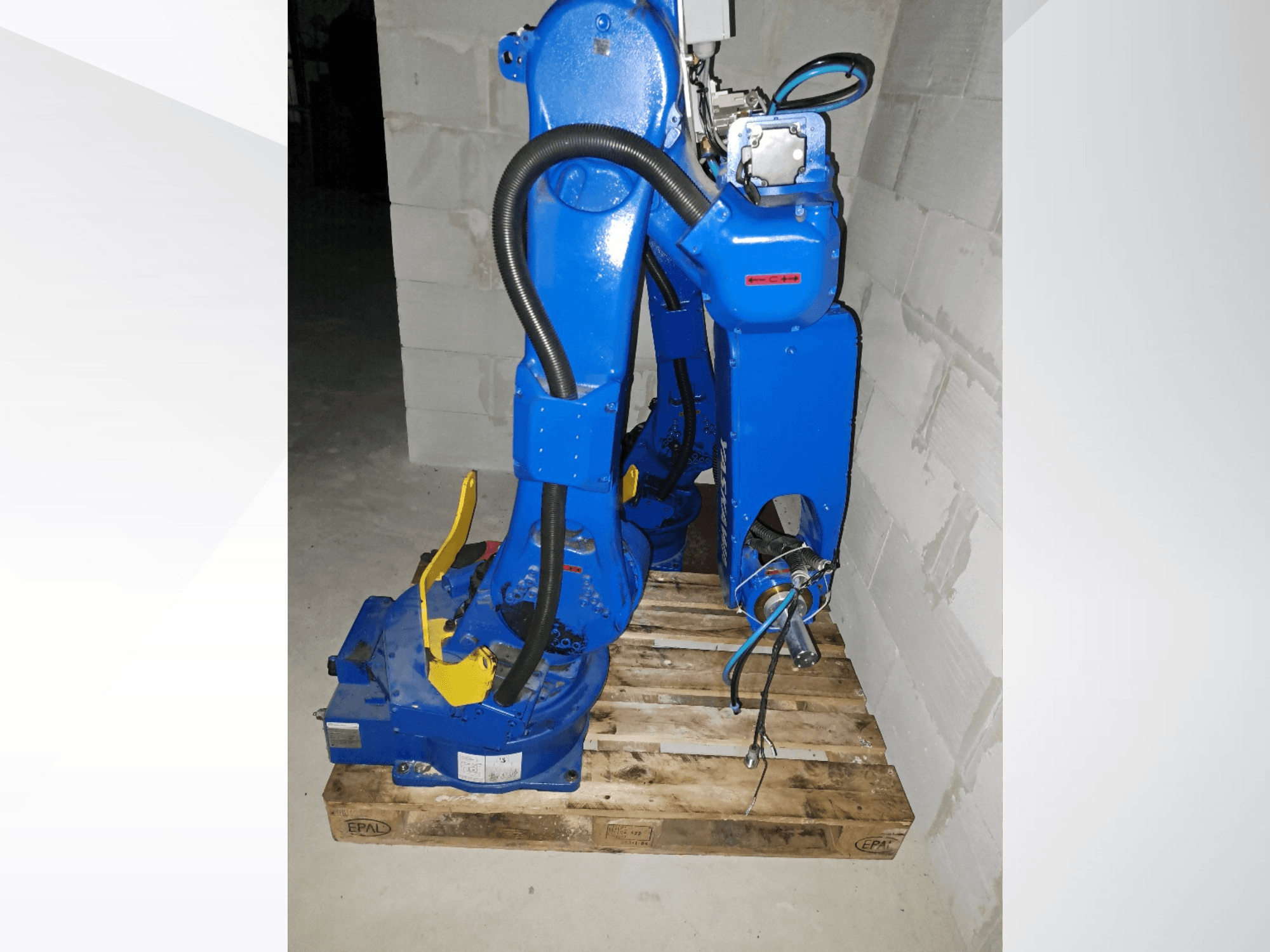 Zilā krāsā izpildīta „Yaskawa Motoman GP25” robota roka, skats no sāniem uz koka paliktņa, redzami vadi un mehāniskie savienojumi.