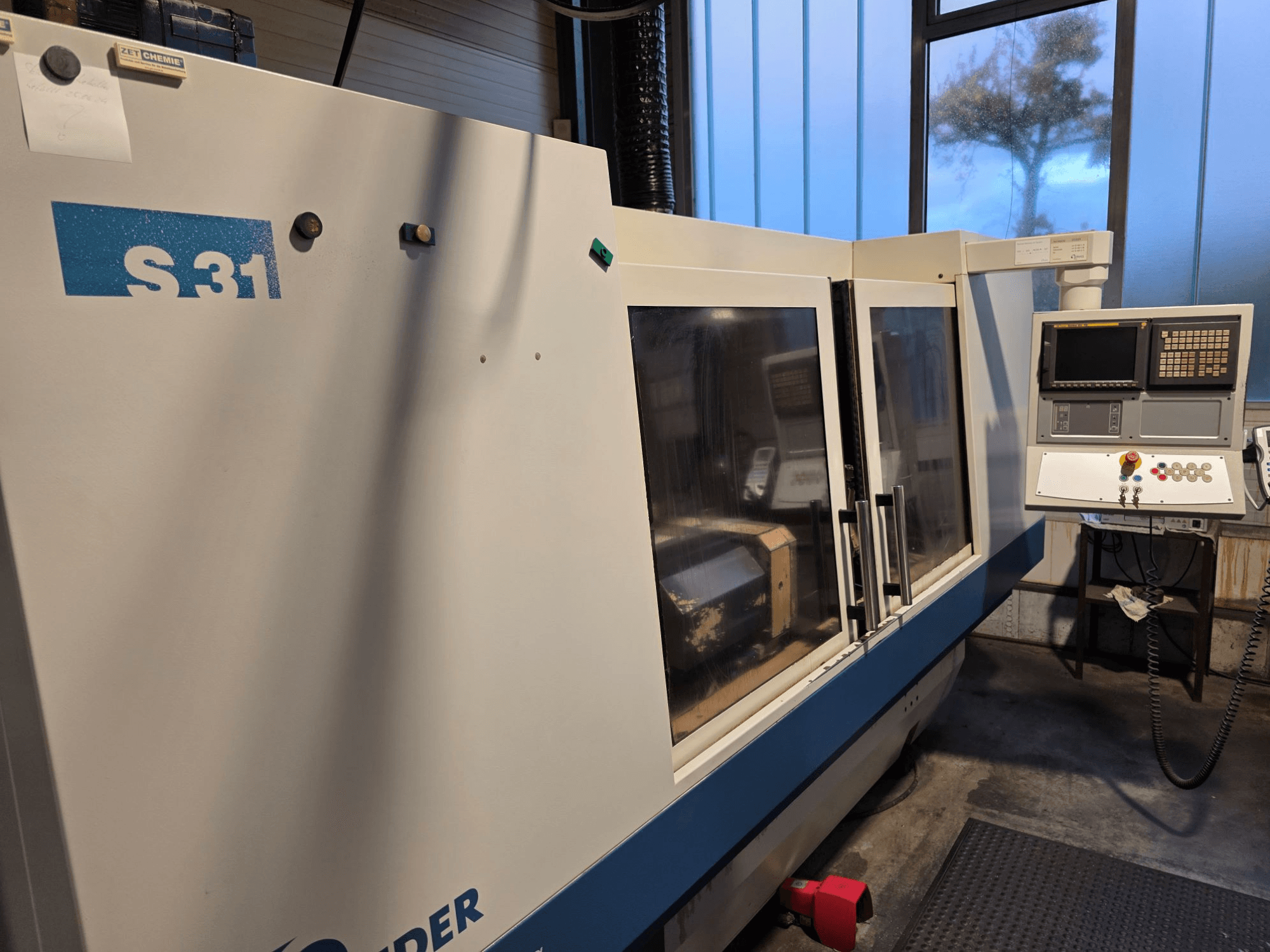 S31 CNC slīpmašīna, skats no priekšpuses, vadības panelis ar ekrānu un pogām, lielas stikla durvis, industriāla vide.