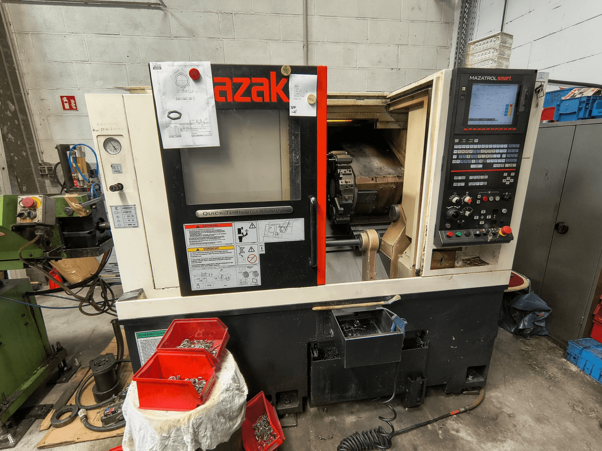 Mazak Quick Turn Smart 100 S, sānskats, kurā redzams vadības panelis, dzesēšanas šķidruma paliktnis un uzglabāšanas kastes ar skrūvēm.