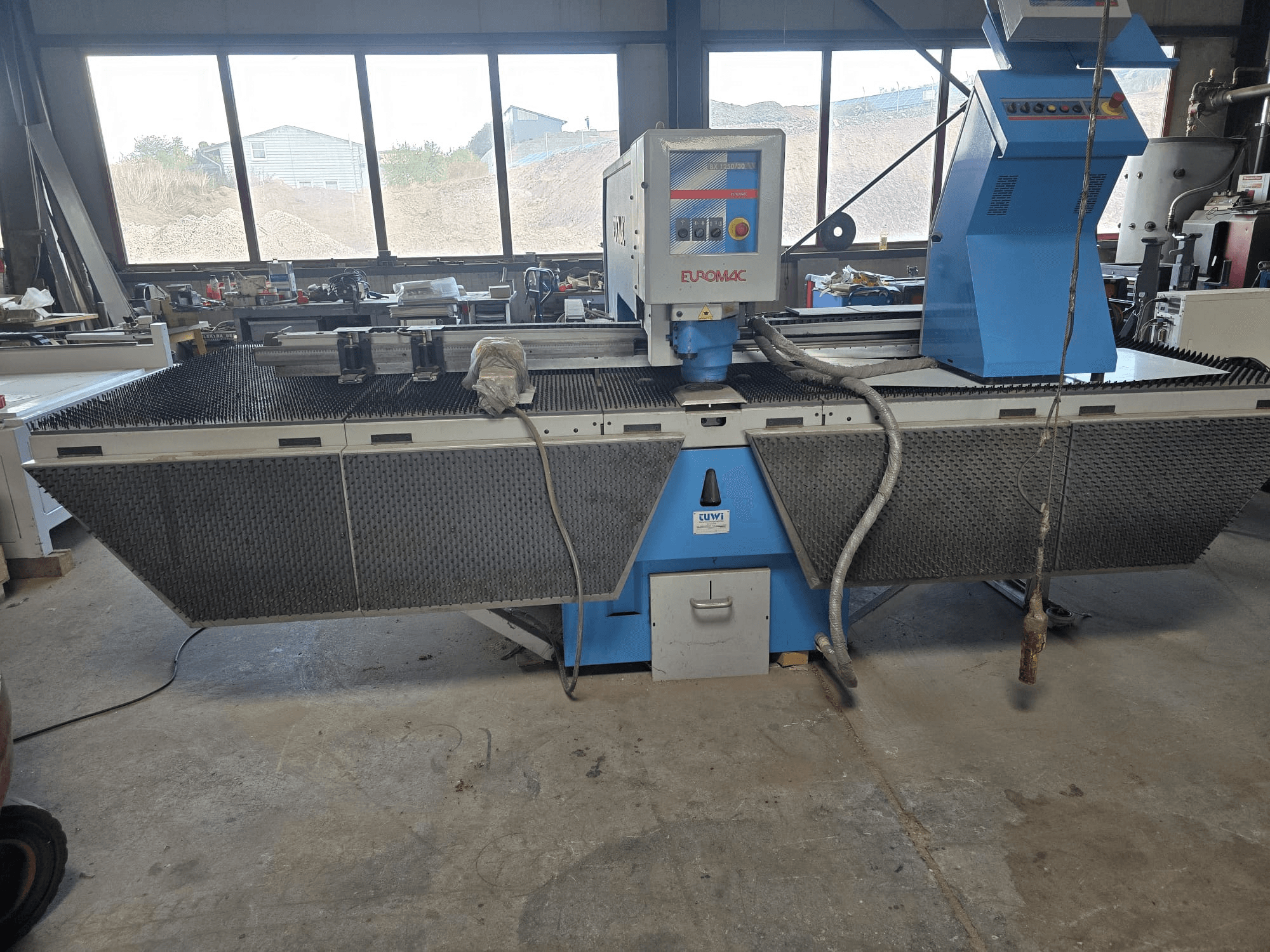 CNC perforācijas mašīna „Euromac BX 1250/30” darbnīcā, sānskats, kurā redzamas instrumentu vadības ierīces, rūpnieciskā dizaina elementi un metāla griešanas galds.