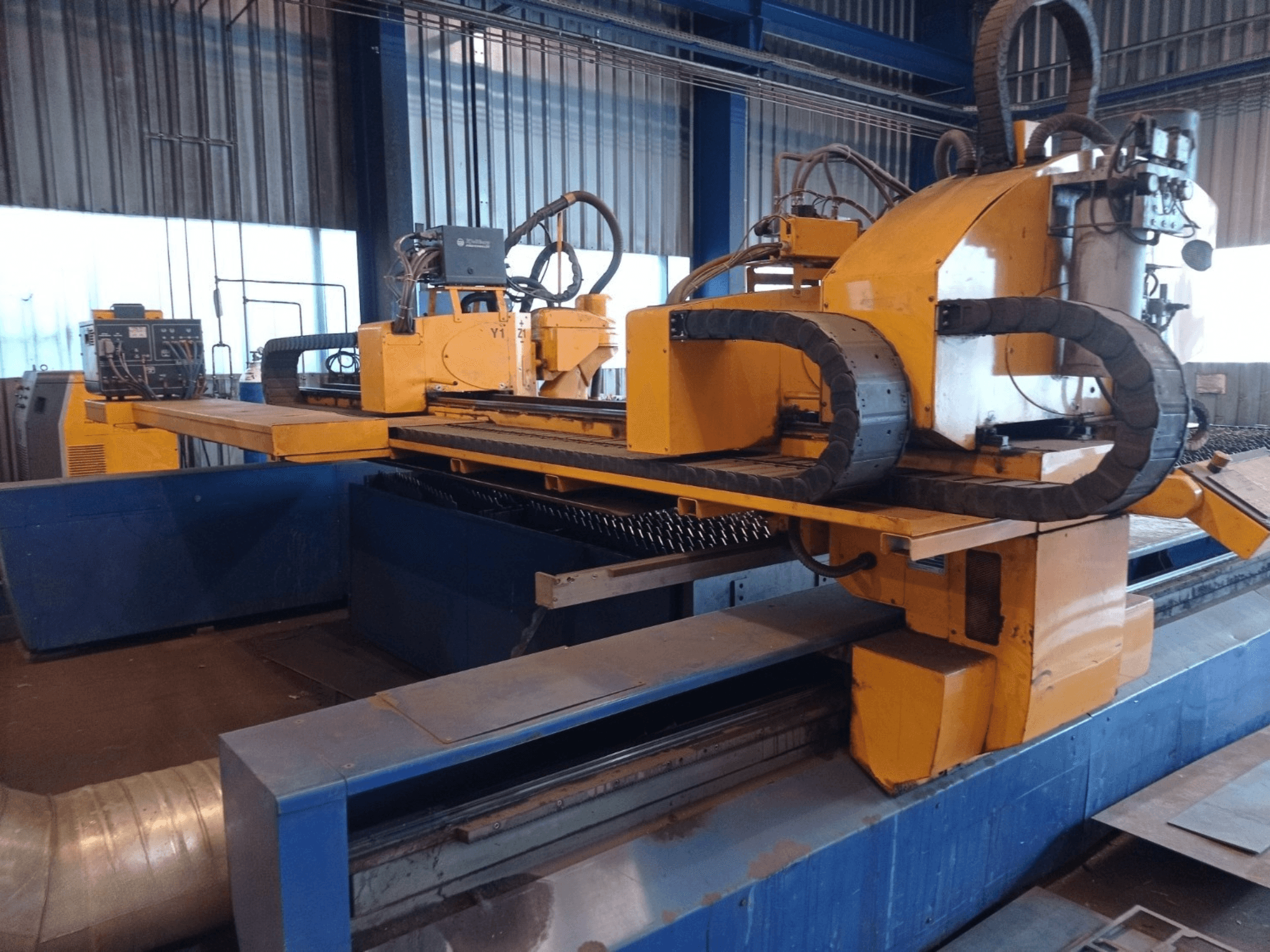 Dzeltena CNC plazmas griešanas iekārta ar divām horizontālām rokām, parādīta no priekšpuses, ar vadības paneli un griešanas gultni.