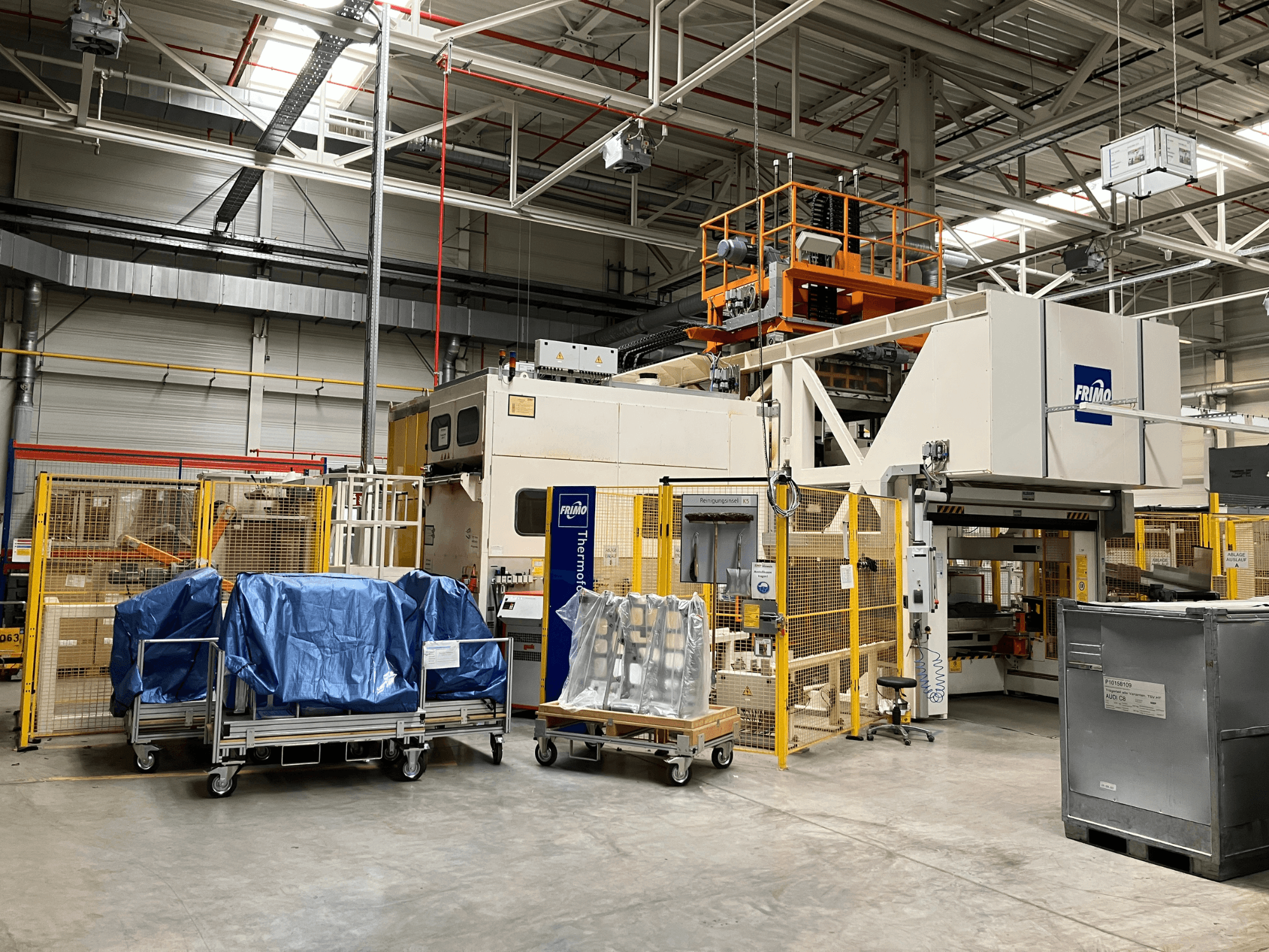 FRIMO FTL 80/200 Fullflex plastmasas apstrādes iekārta rūpnīcā, skats no priekšpuses ar redzamiem ziliem brezentiem un drošības barjerām.