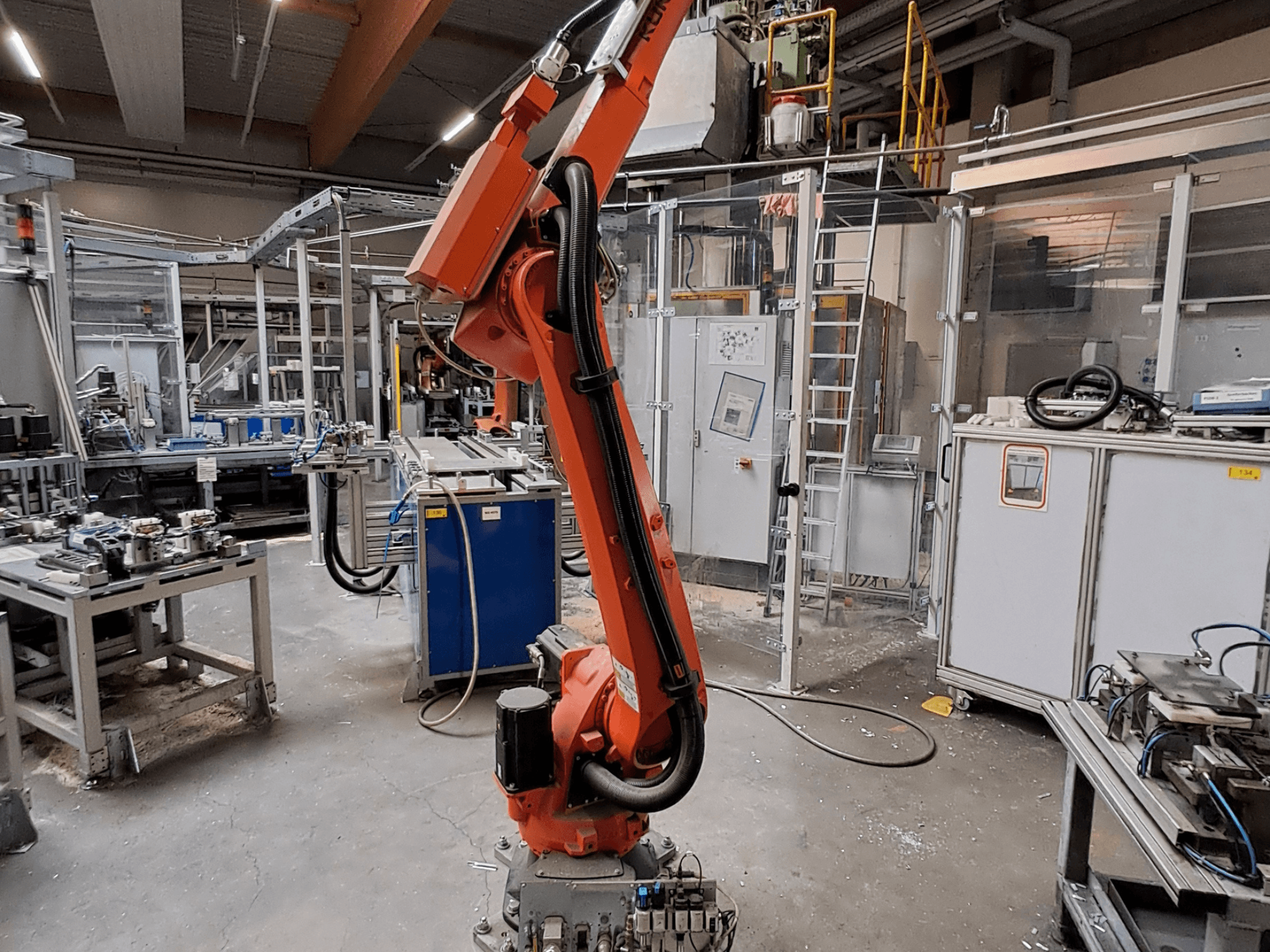 KUKA KR 26 R3100/SEL robota roka rūpnieciskā vidē, redzama no sāniem, fonā redzams darba aprīkojums.