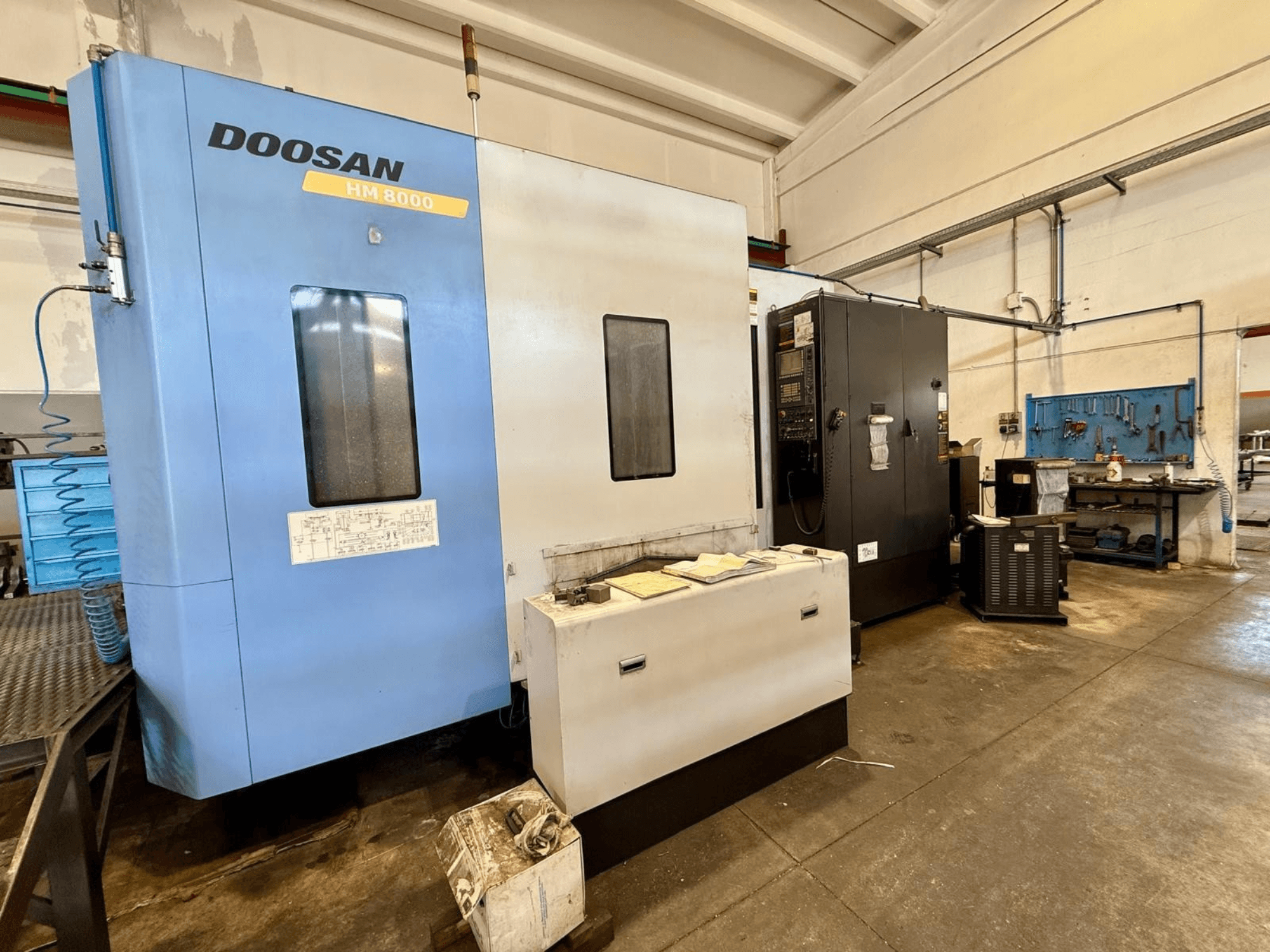 Zilā krāsā izpildītais horizontālais apstrādes centrs „DOOSAN HM 8000“ — skats no priekšpuses, kurā redzams vadības panelis un blakus esošais darbnīcas aprīkojums.
