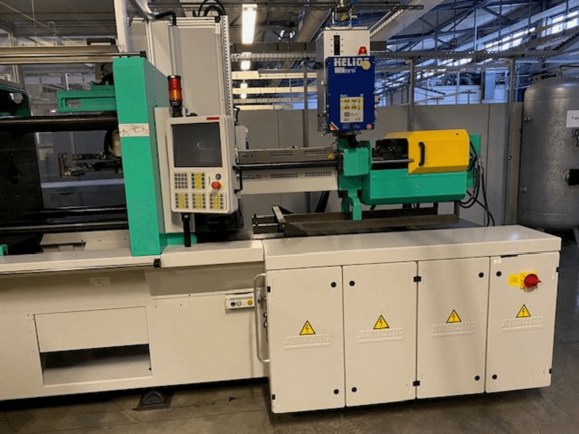 Rūpnieciskās liešanas mašīnas, HELIO modelis, skats no priekšpuses, ar vadības paneli, drošības aizsargiem un avārijas apturēšanas pogu.