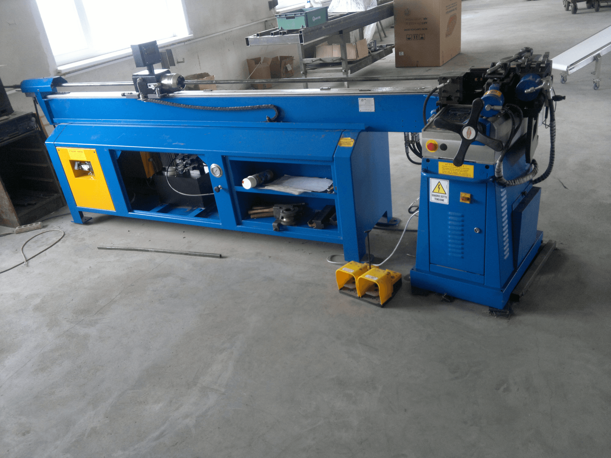 Zila kokapstrādes mašīna ar dzelteniem akcentiem, skats no sāniem; vadības panelis, pedāļi un rotējošs asmens komplekts.