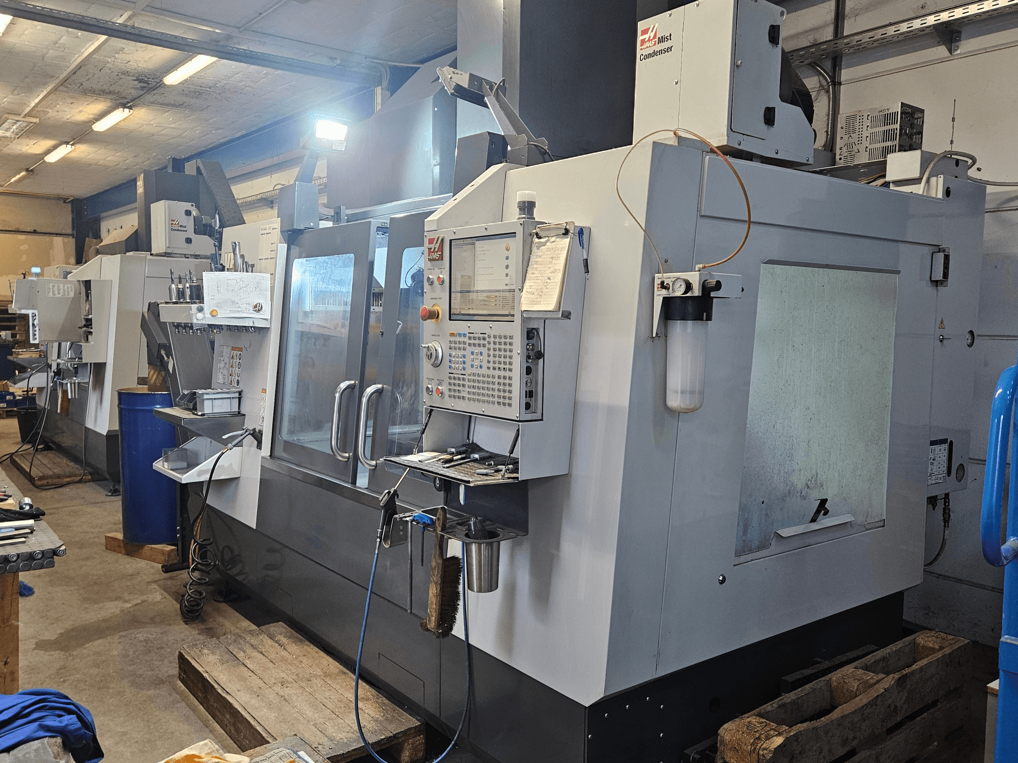 Vertikālais apstrādes centrs „Haas VF-3YT/50“ darbnīcā — skats no priekšpuses, kurā redzams vadības panelis, instrumentu turētāji un miglas kondensators.