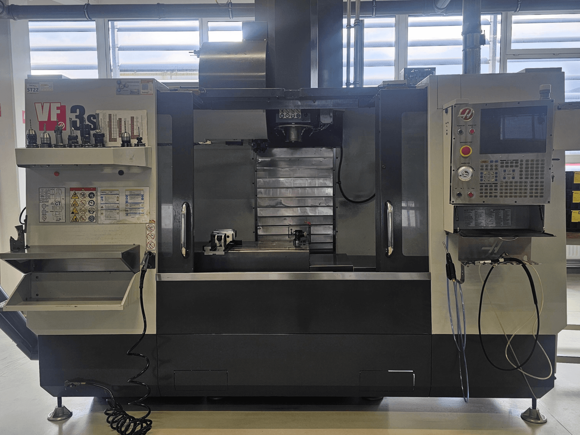 Haas VF3SS CNC frēze, skats no priekšpuses, ar instrumentu turētājiem, vadības paneli un tukšu dzesēšanas šķidruma paplāti, ar redzamiem drošības marķējumiem.