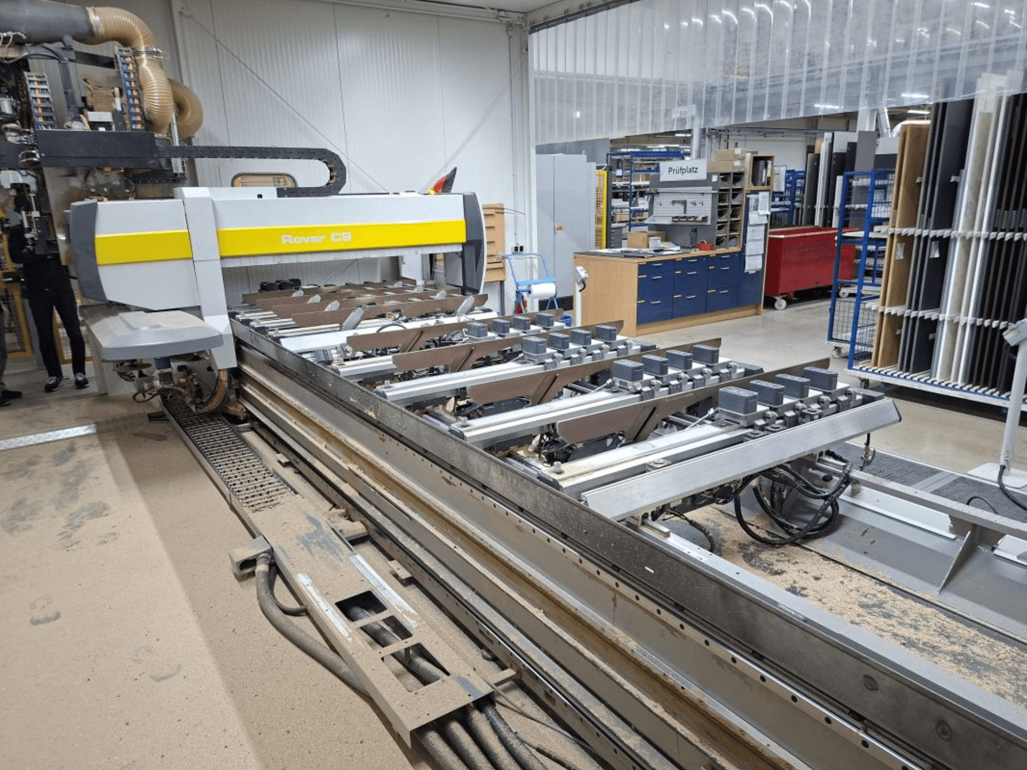 Rover C9 CNC mašīna darbnīcā; priekšpusē redzami griezējinstrumenti un konveijera sistēma, uz grīdas sakrājušies putekļi.