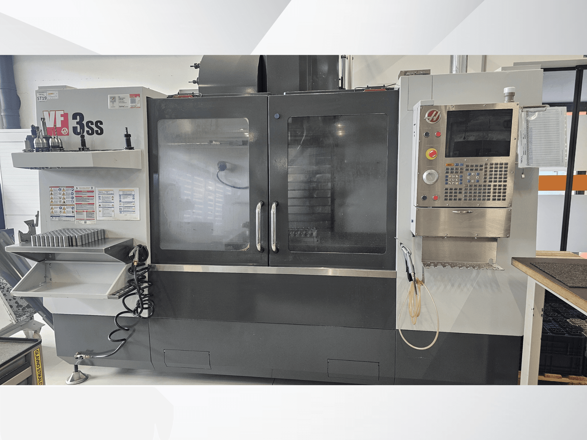 HAAS VF-3SS vertikālais apstrādes centrs, skats no priekšpuses, ar vadības paneli, instrumentu plauktu un lielām stikla durvīm.