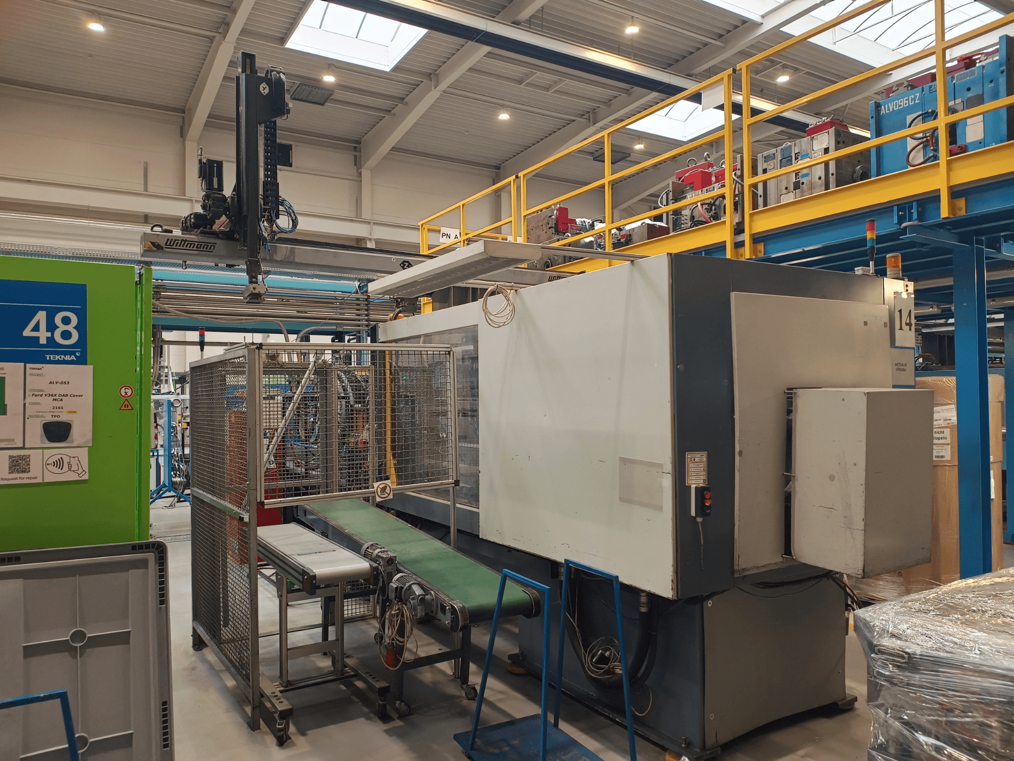 Winmon rūpnieciskās automatizācijas iekārta, sānu skats, kurā redzama konveijera lentes, vadības panelis un drošības būris rūpnīcā.