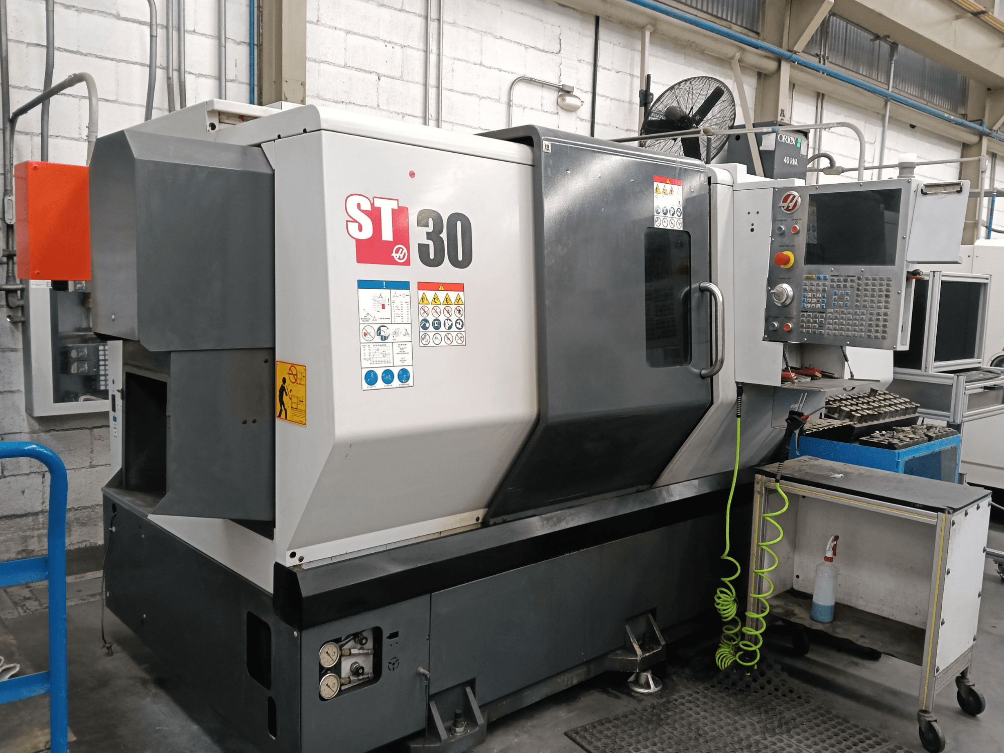 Alternatīvais teksts: HAAS ST-30 horizontālās virpošanas mašīnas skats no priekšpuses ar vadības paneli, drošības uzlīmēm un instrumentiem blakus esošajā ratiņos.