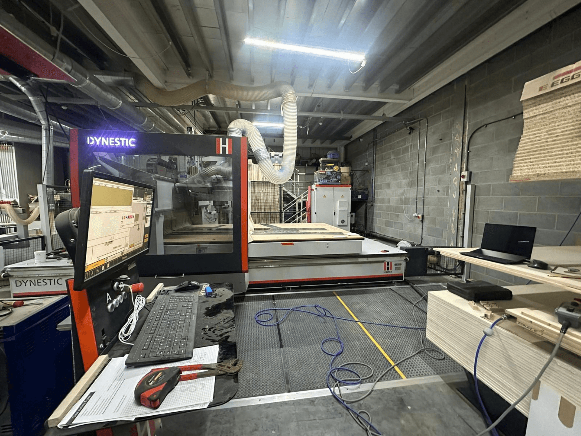 HOLZHER Dynestic 7535 koka frēze darbnīcā, vadības panelis, monitors un materiāli uz darba galda, skats no priekšpuses.