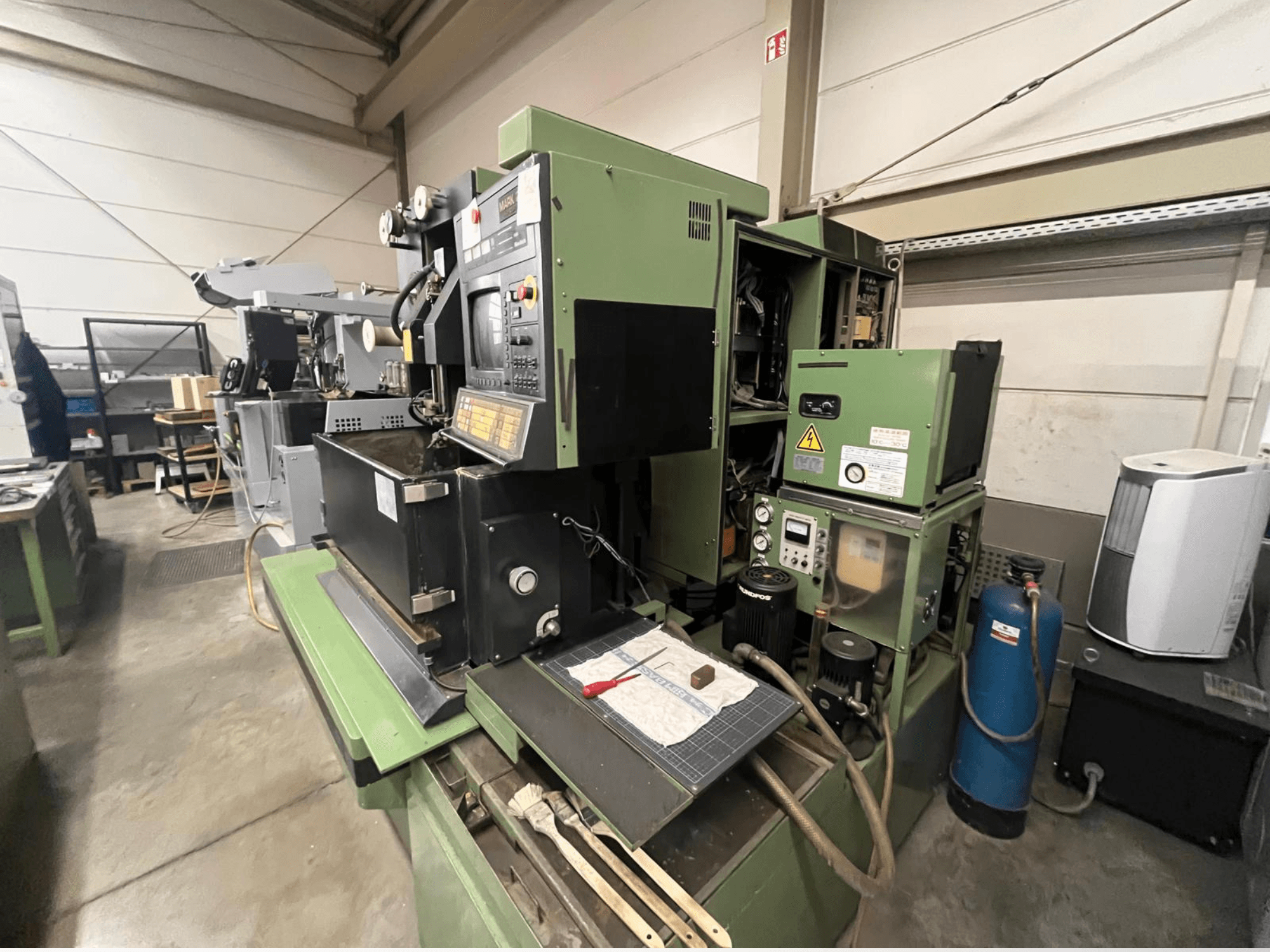 Zaļa industriālā mašīna ar vadības paneli, skats no sāniem; ietver hidrauliskos komponentus un darba virsmas laukumu ar instrumentiem.