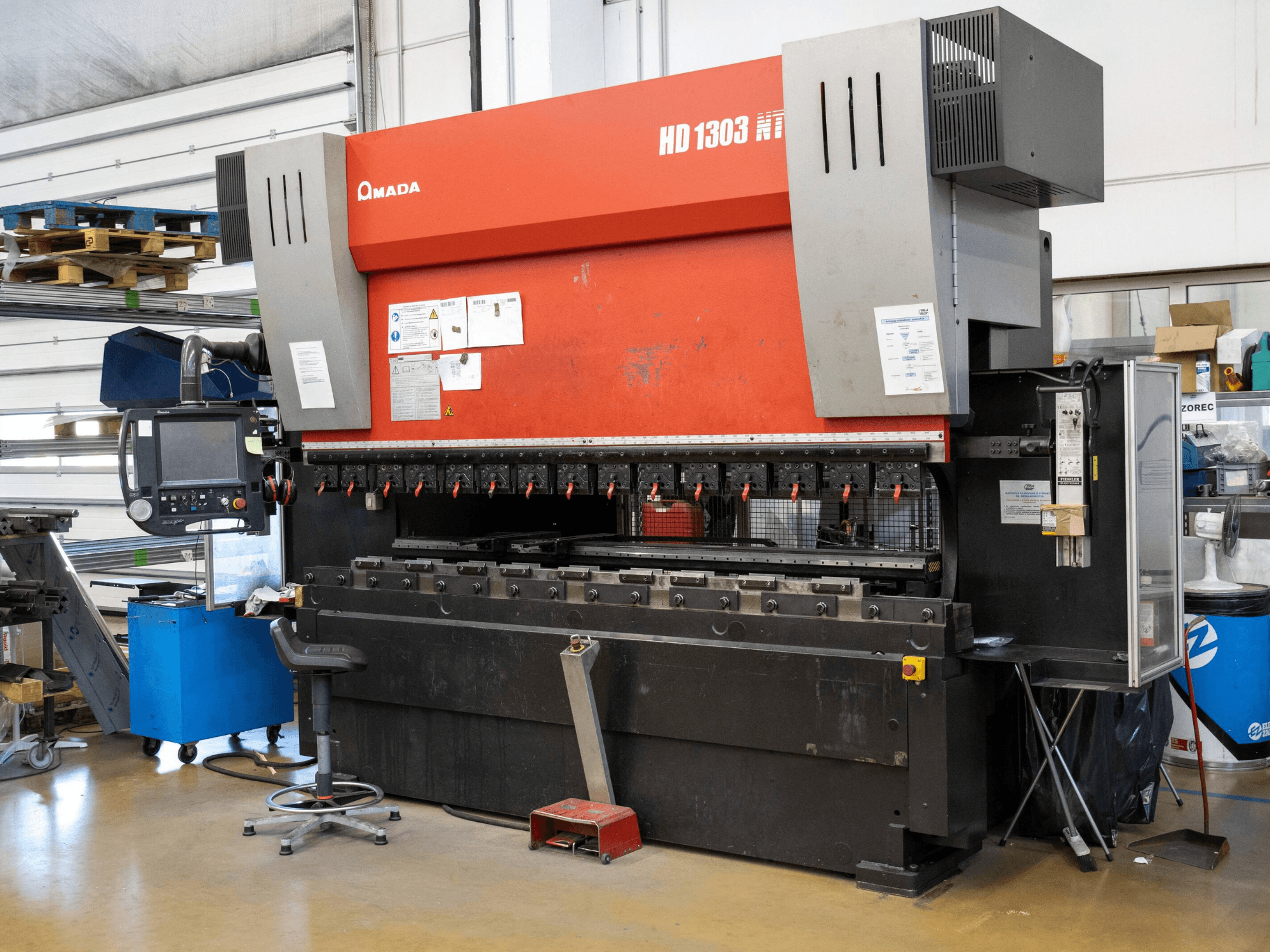 Amada HD 1303 hidrauliskā preses bremze darbnīcā, skats no priekšpuses; ar vairākām skavām, vadības paneli un regulējamu krēslu.