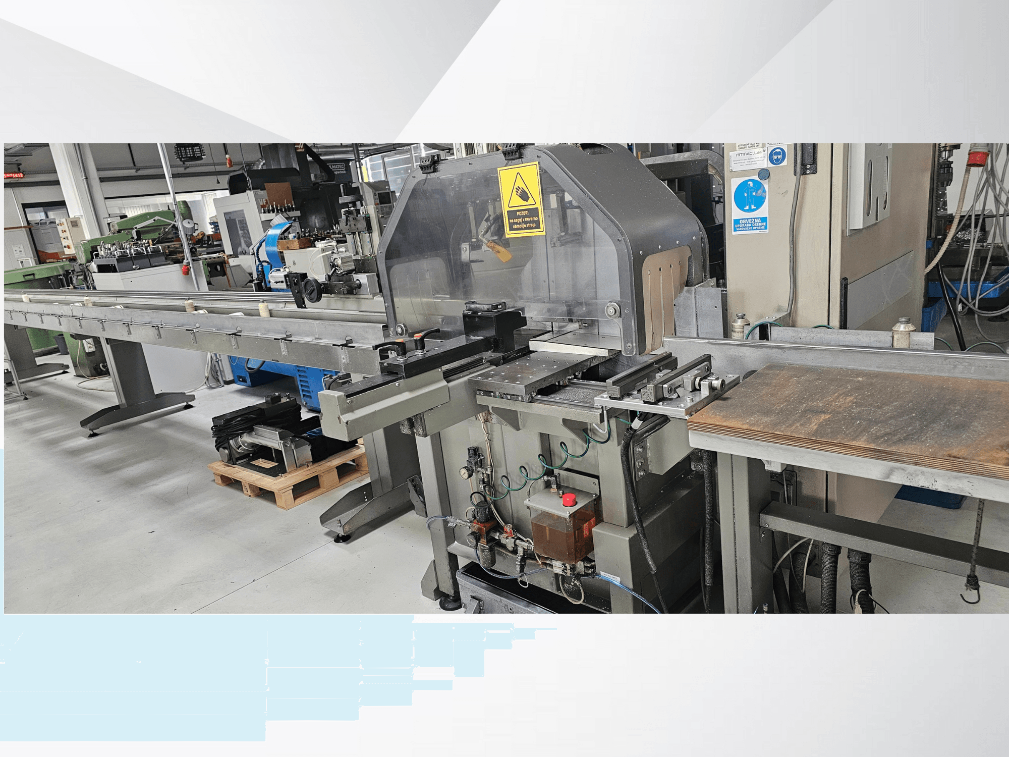 Rūpnieciskā griešanas iekārta no sānu skata, ar garu konveijeru, vadības paneli un drošības brīdinājumiem.