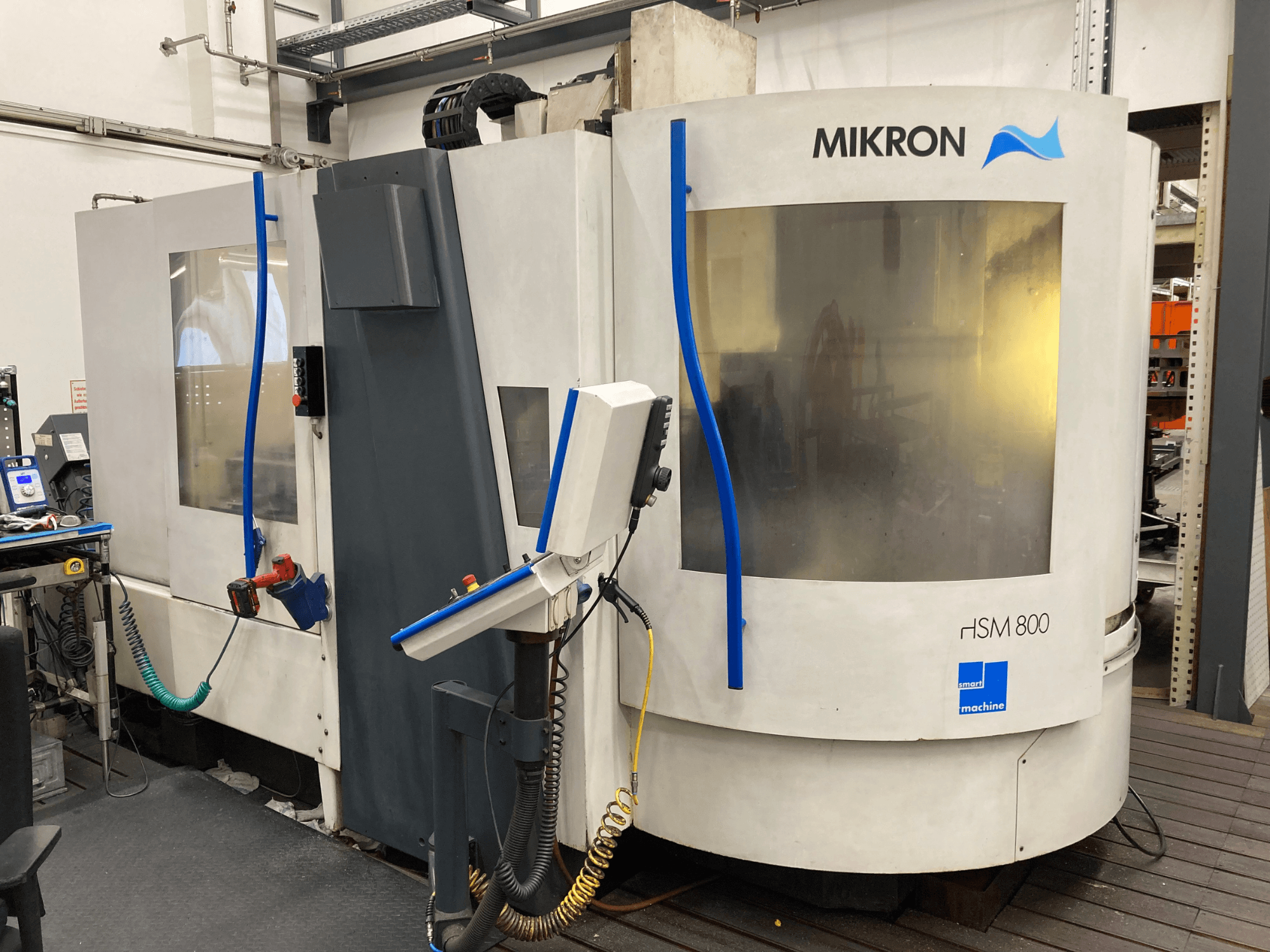 Mikron HSM 800, CNC apstrādes centra sānu skats ar vadības paneli un instrumentu turētāju, redzamas šļūtenes un kabeļi.