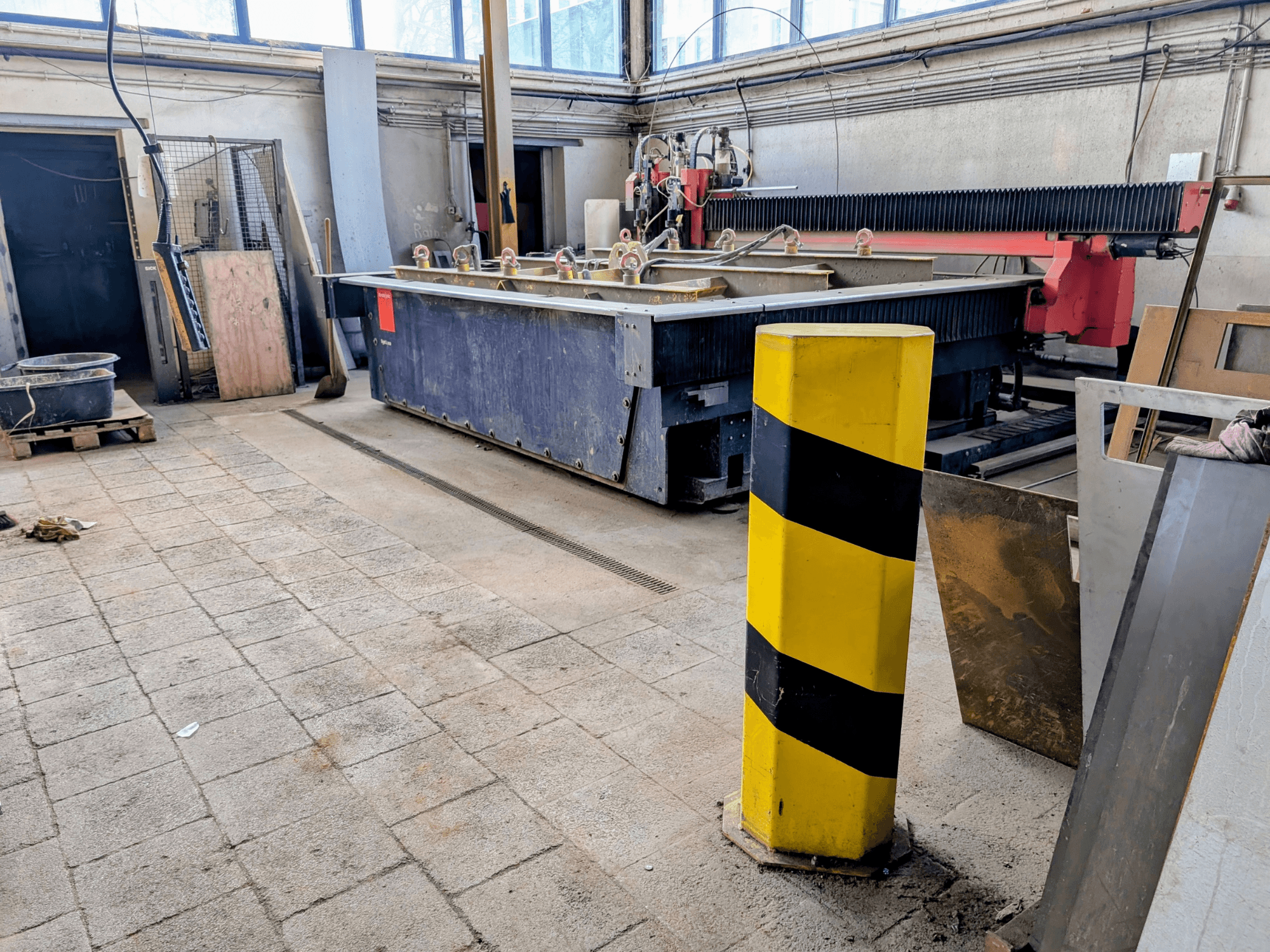 Bystronic Byjet L 2030 ūdensstrūklas griešanas iekārta darbnīcā, skats no sāniem, ar dzeltenu drošības stabu un apkārtējiem materiāliem.