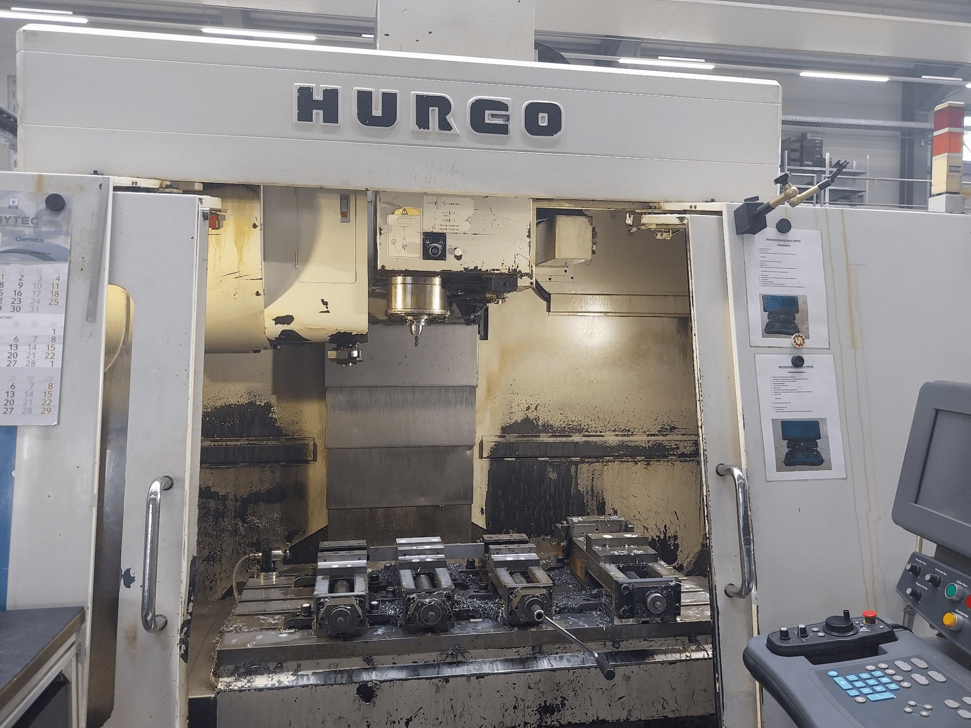 Hurco VMX 42 vertikālais apstrādes centrs priekšpusē, redzams darba galds ar piederumiem un vadības panelis, nedaudz nolietots.